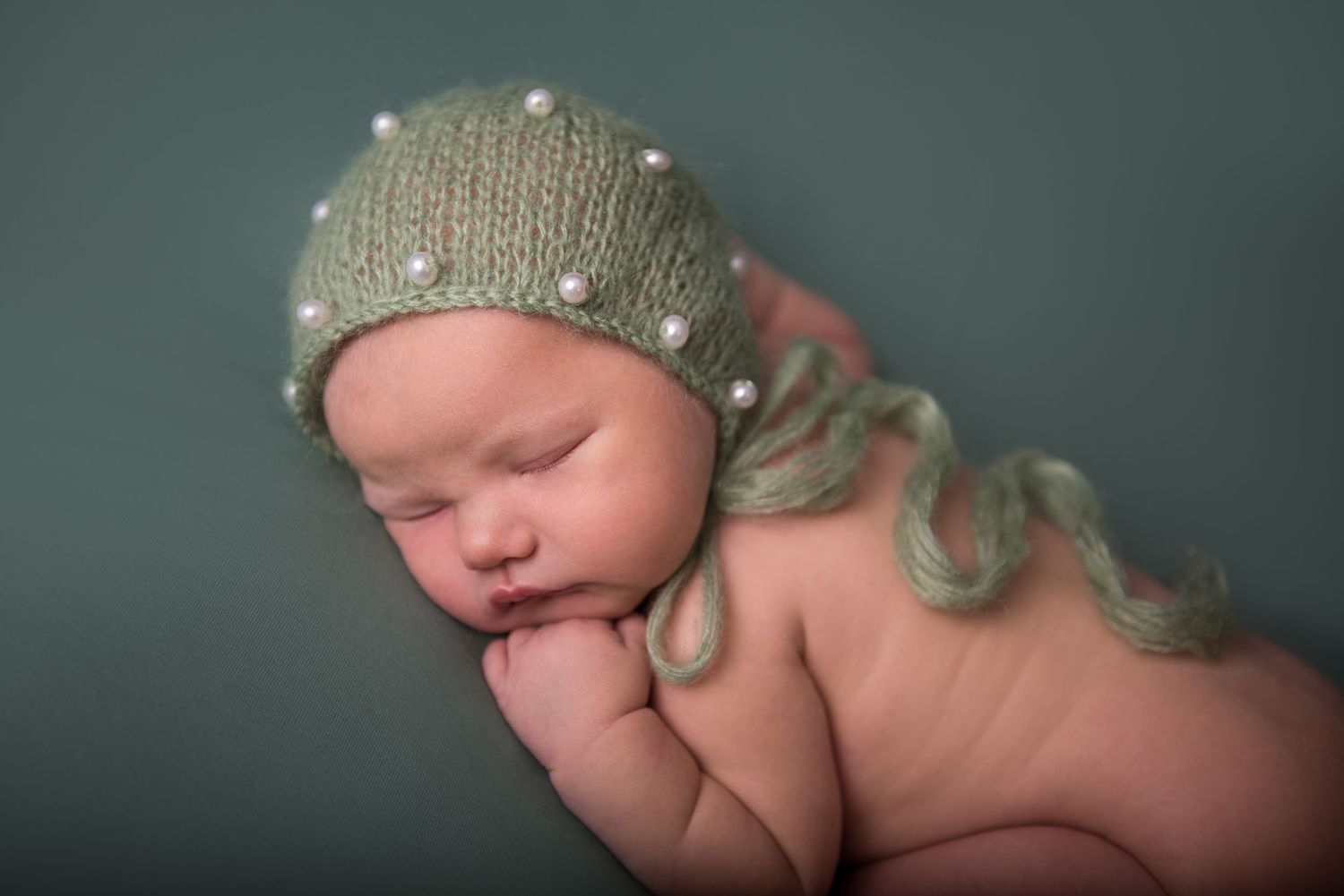 Peaceful sleeping newborn wearing a soft knitted beanie in a gentle green-toned studio setting.