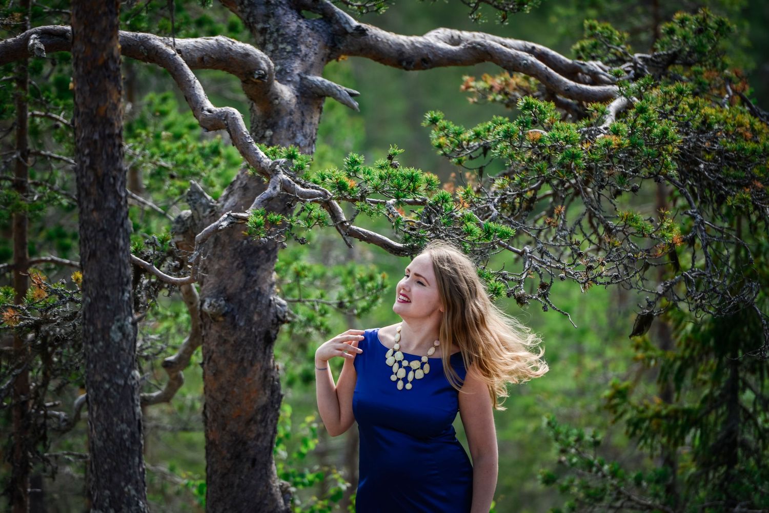 A young woman in a blue dress laughs joyfully among lush green forest trees.