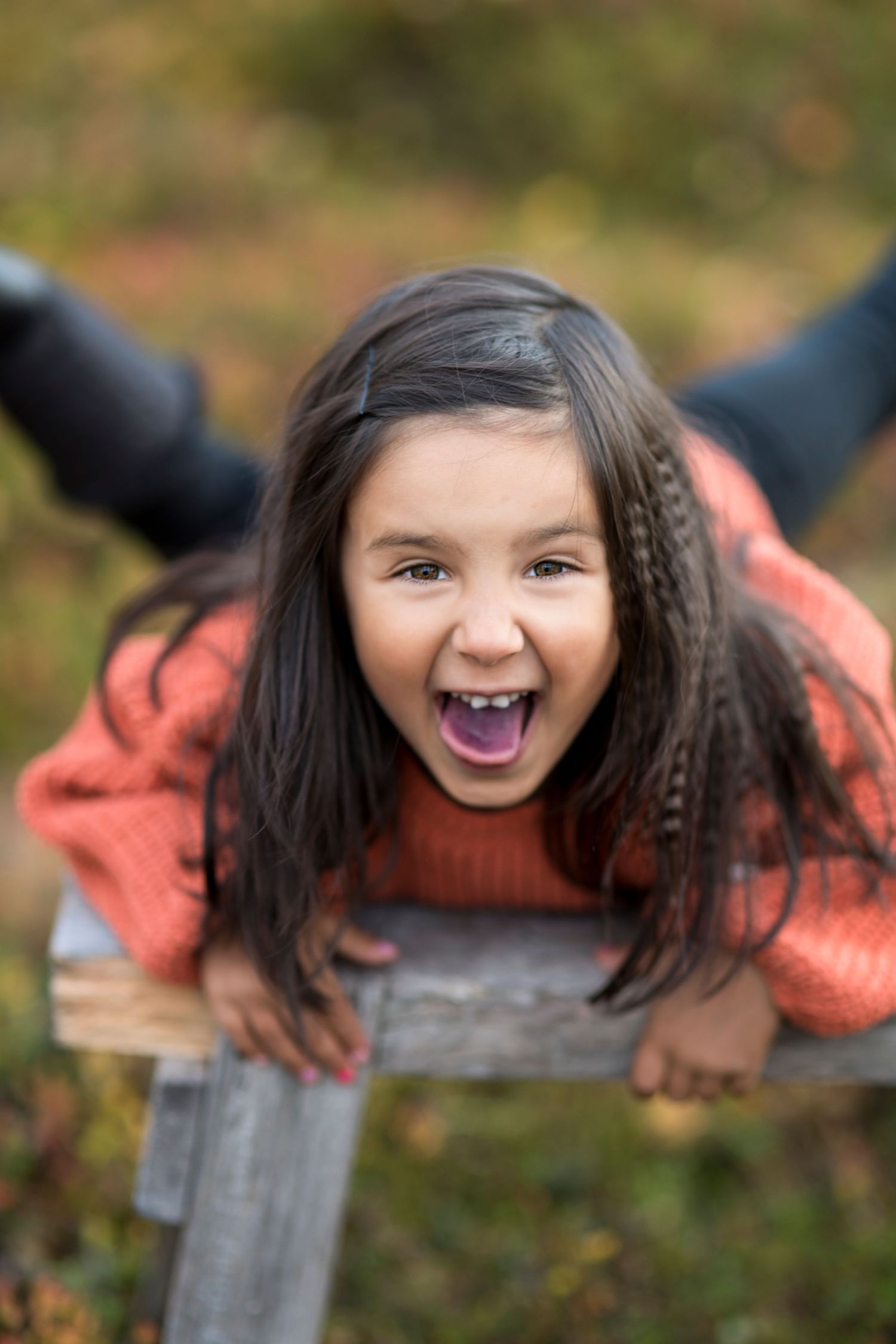 Excited young girl in orange sweater leaning on wooden fence outdoors, laughing with mouth open.