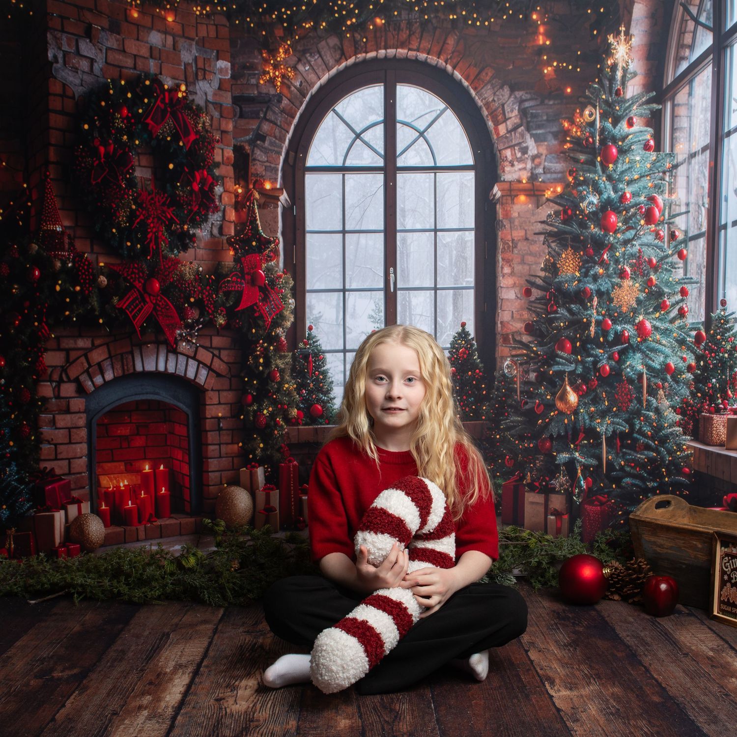 A young girl in a red sweater sits cross-legged holding a candy cane sock beside a lit fireplace and Christmas tree.