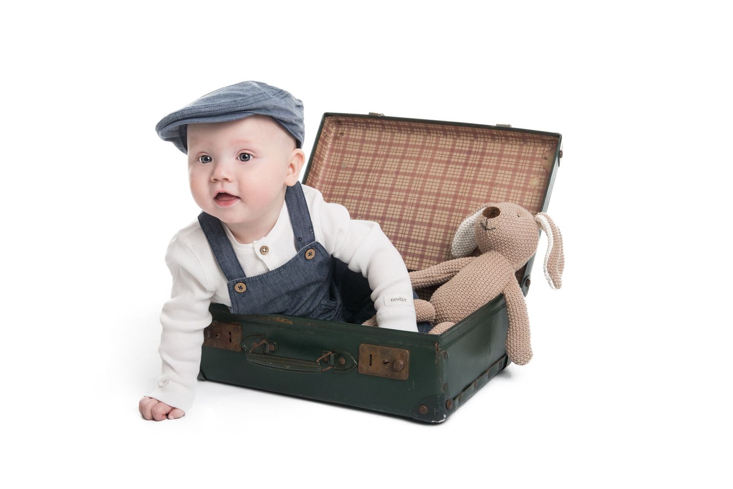 Baby in vintage outfit playing with antique suitcase on white backdrop.