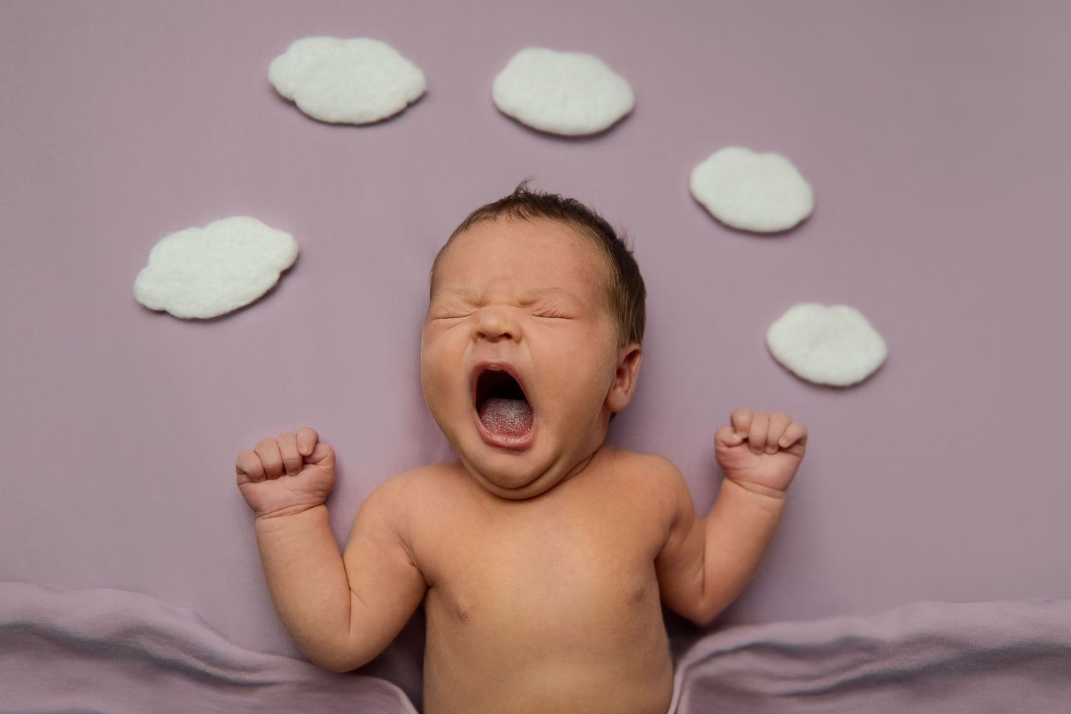 Newborn baby sleeping on pink backdrop with decorative cloud cutouts.