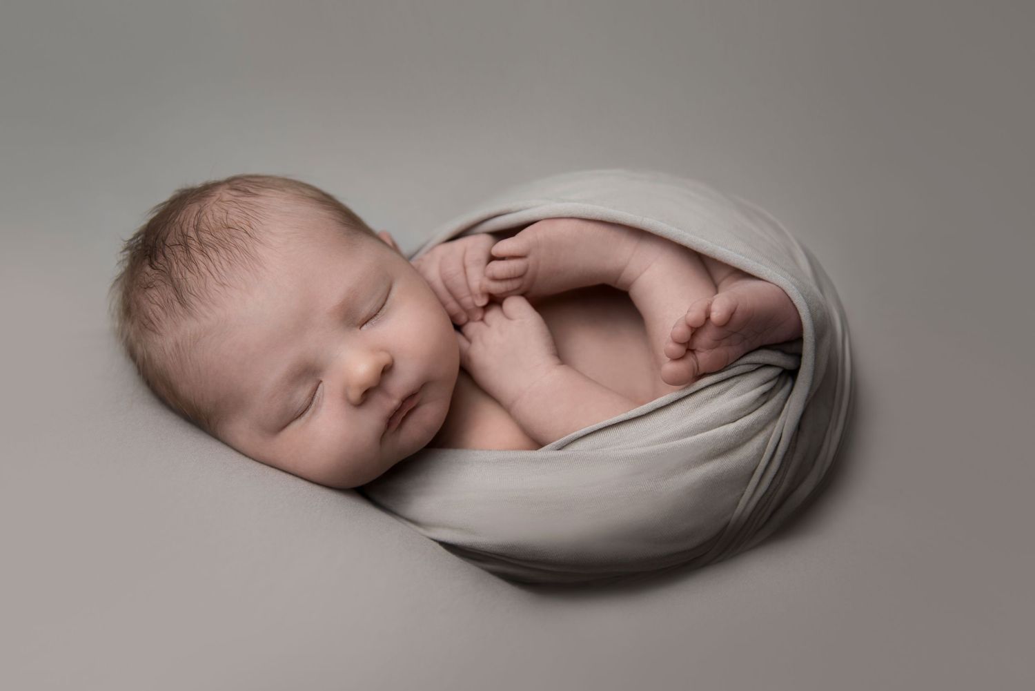 Sleeping newborn wrapped in soft gray fabric in minimalist studio portrait.