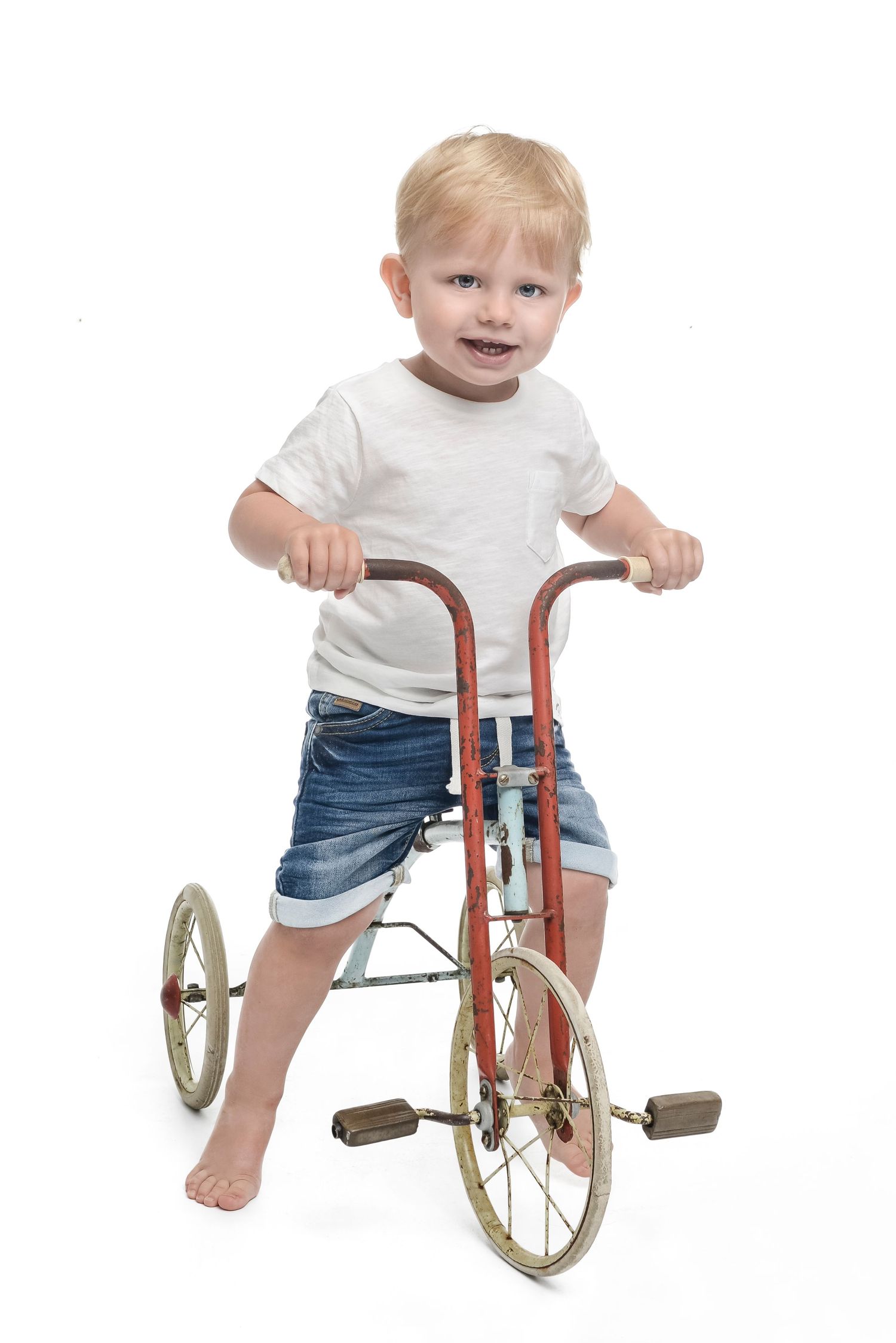 A toddler in white shirt and denim shorts plays with a vintage tricycle.