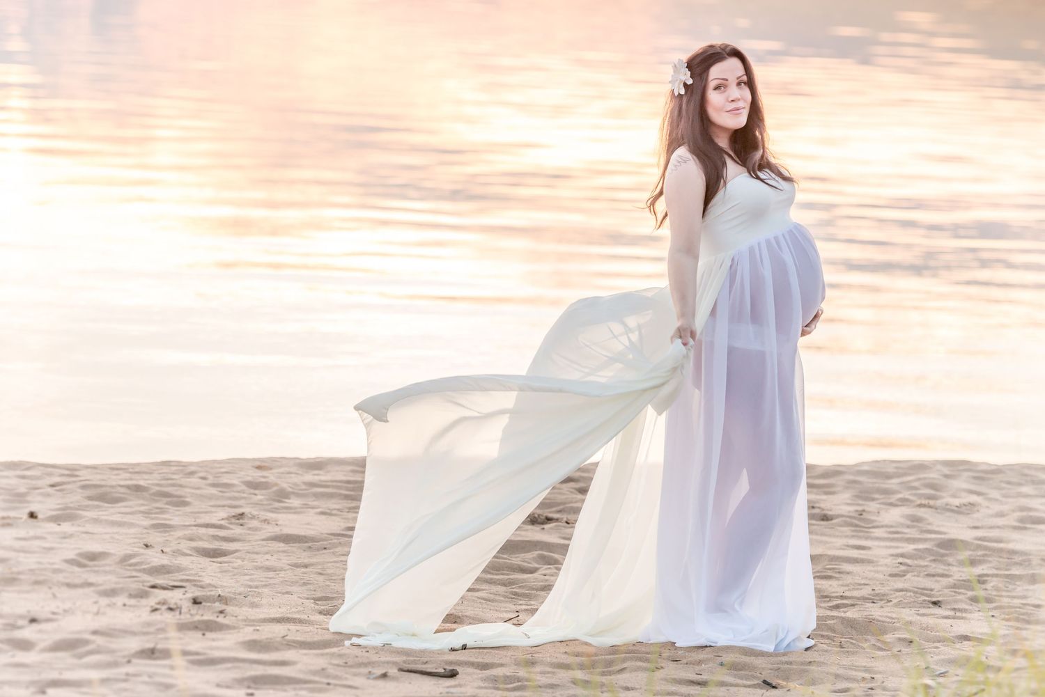 Pregnant woman in flowing white dress standing on sandy beach near water during golden hour sunset.