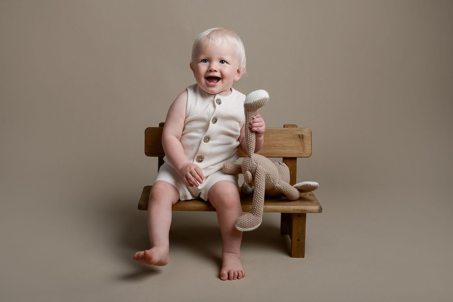 Baby in white romper sits on wooden bench with stuffed toy.