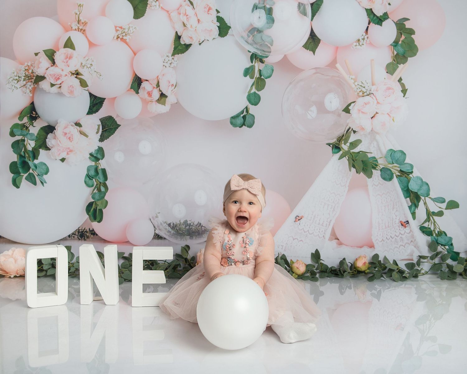 Elegant first birthday setup with pink and white balloons, greenery and decorative letters.