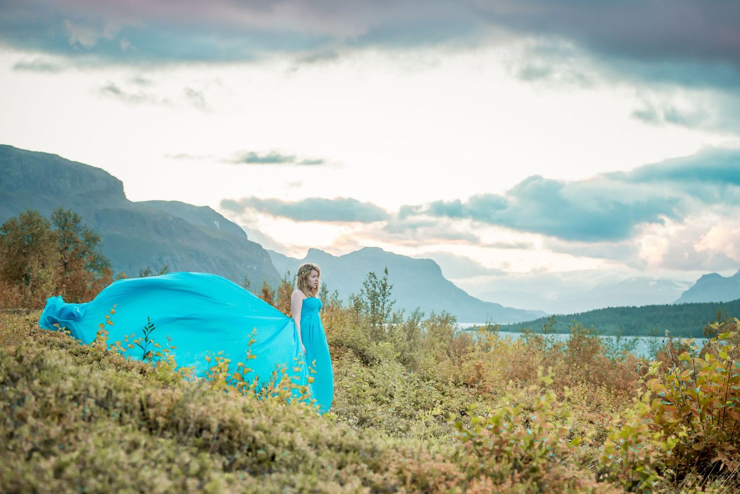Woman in flowing turquoise gown stands amid greenery with mountains and lake backdrop under dramatic cloudy sky.