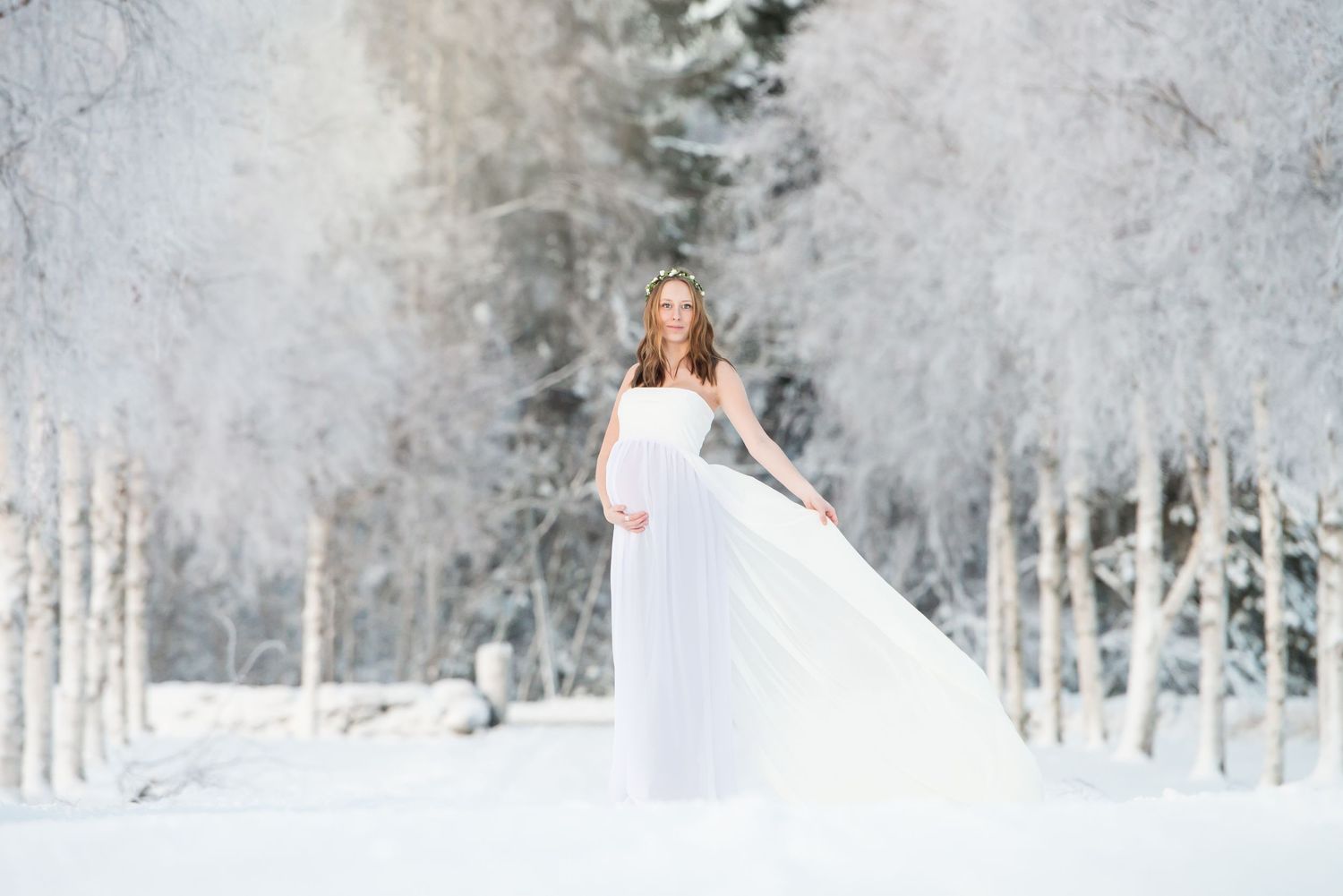 Pregnant woman in flowing white gown standing in frost-covered winter forest for elegant maternity shoot.