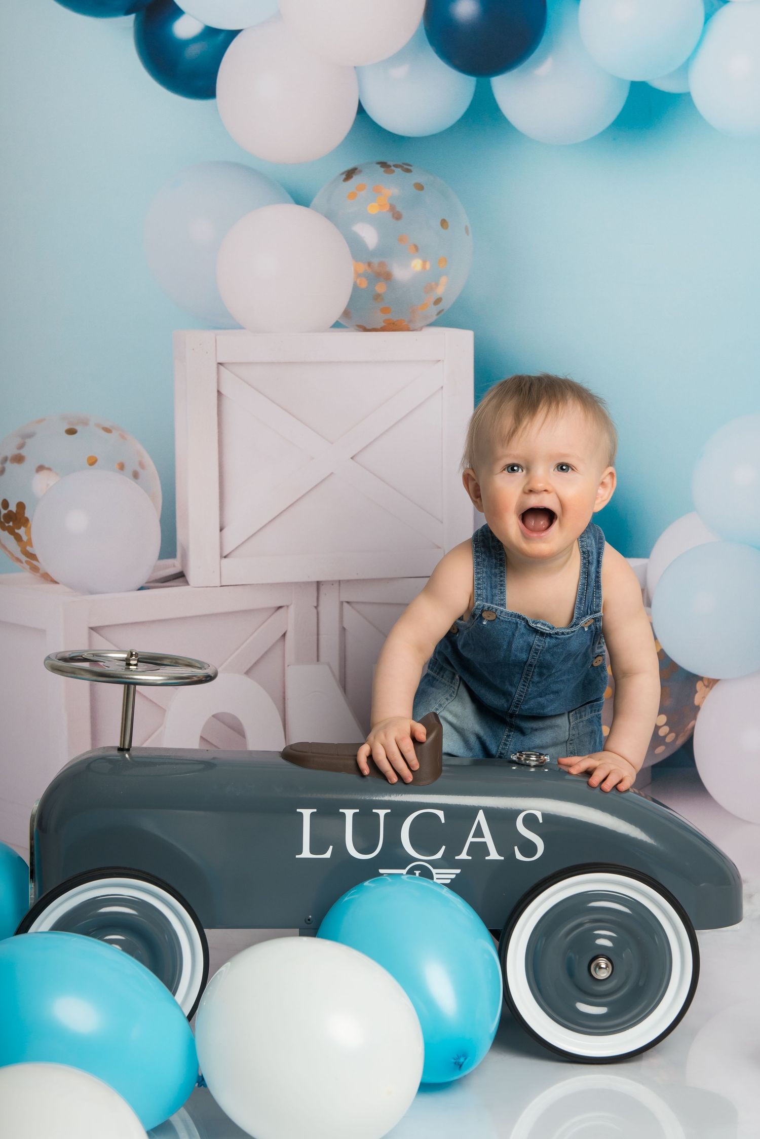 A festive first birthday setup with blue and white balloons, a toy car, and white decorative boxes against a light blue backdrop.