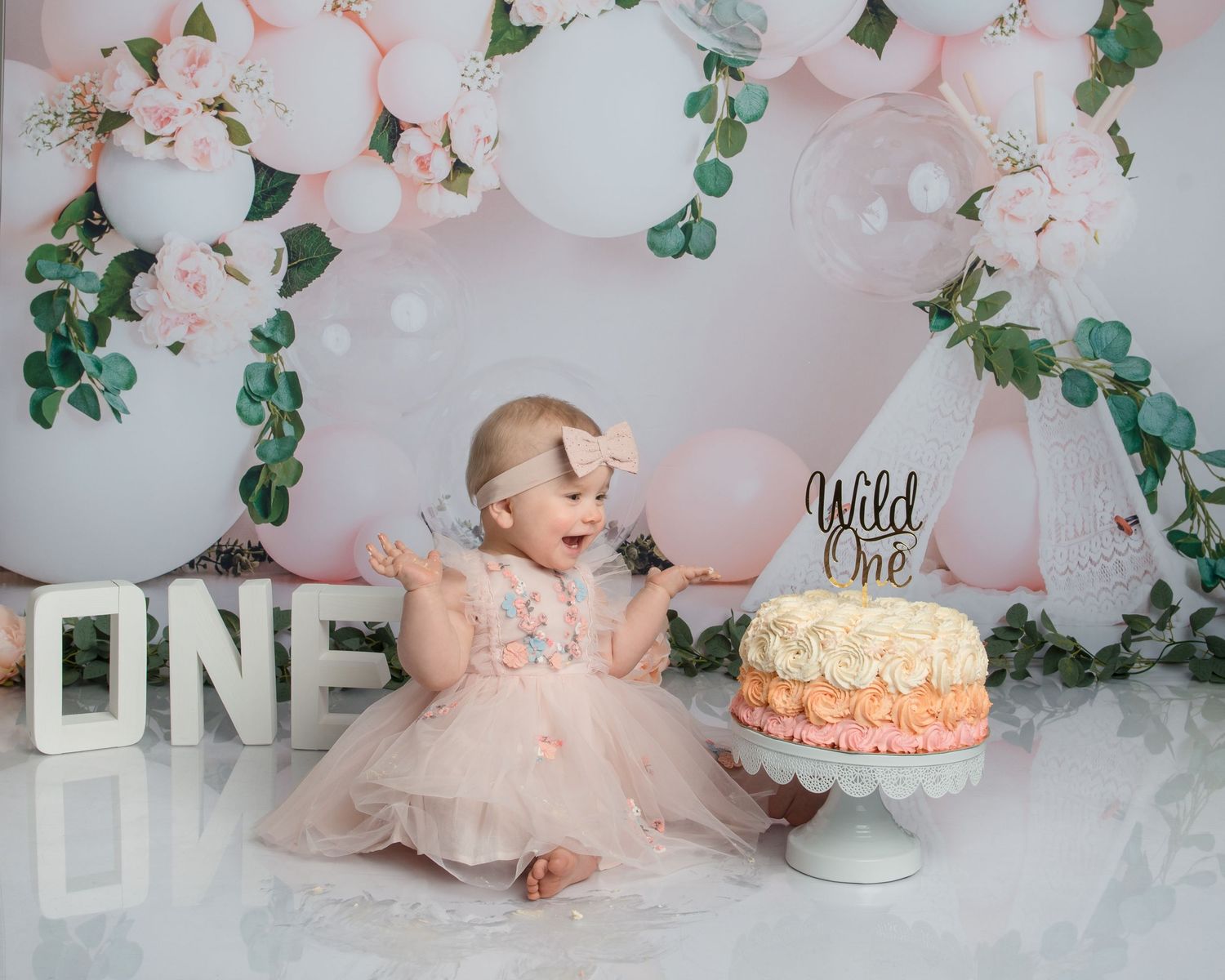 Floral first birthday cake smash setup with pink balloons, greenery and white letters.