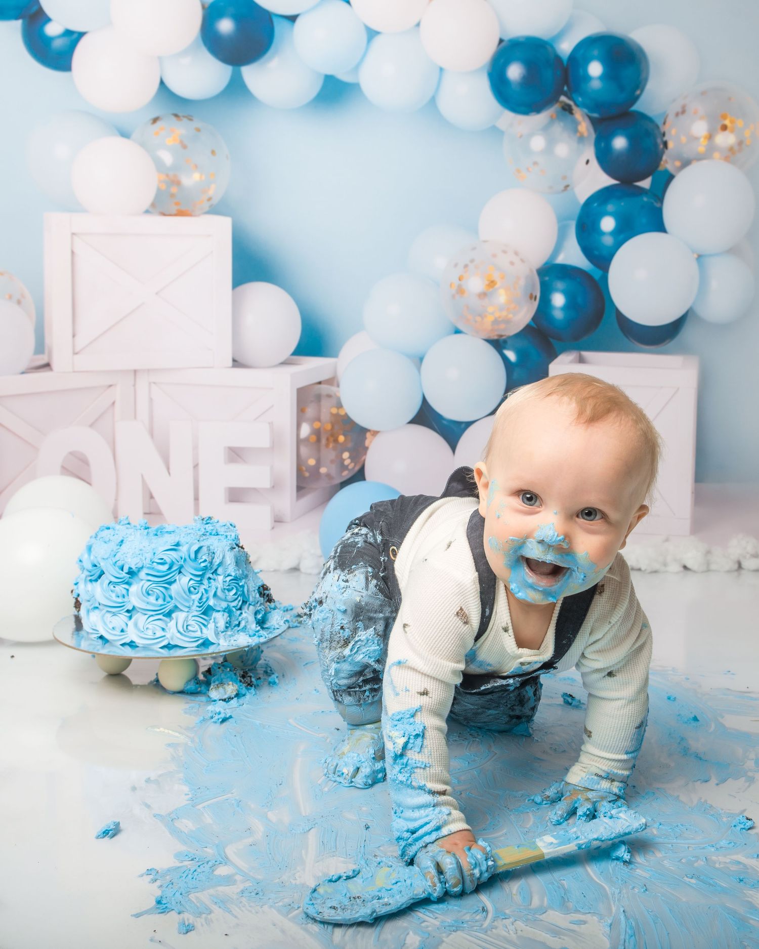 Cake smash photo session with blue and white balloon arch and frosting-covered floor.