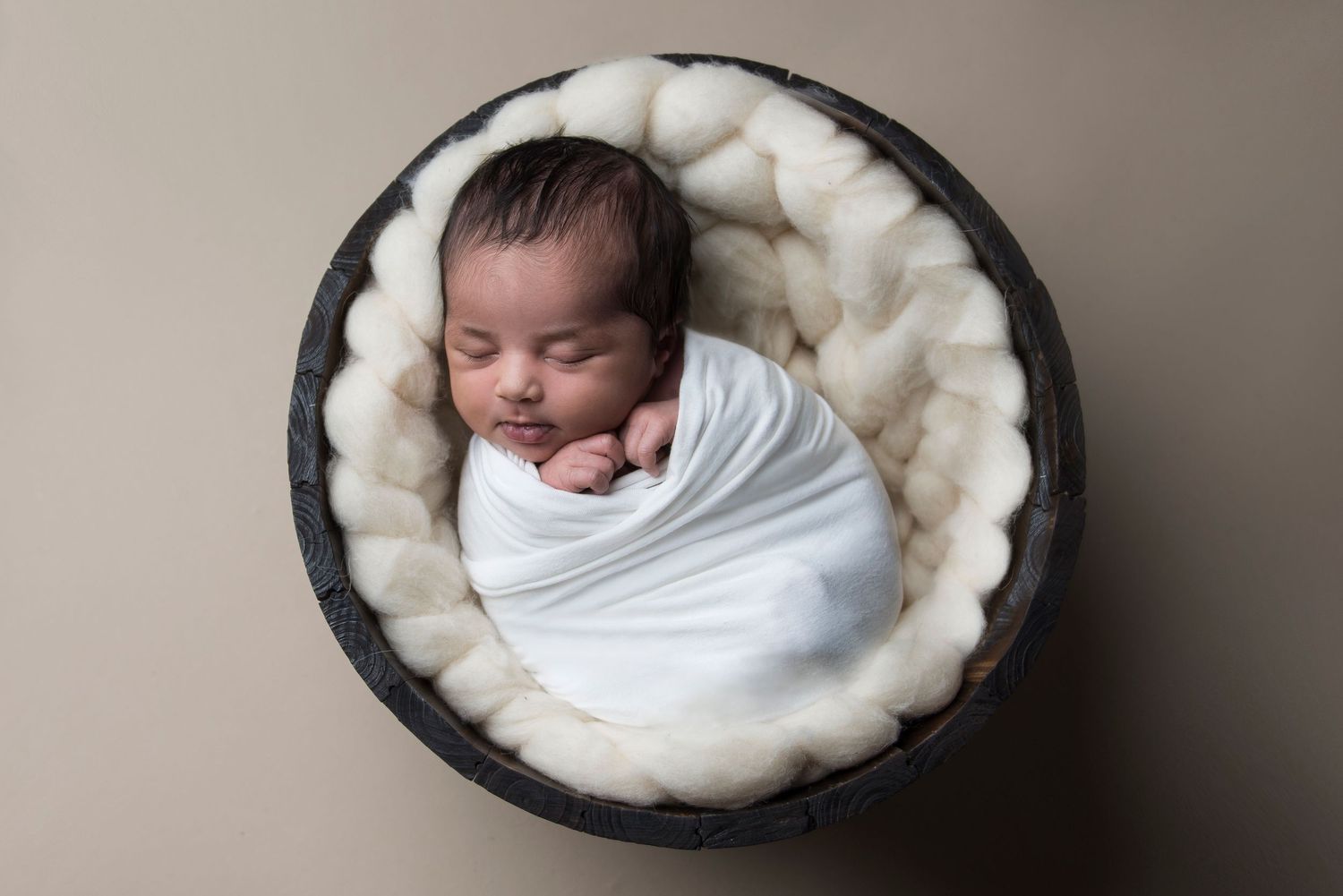 Overhead view of sleeping newborn in white wrap posed in dark round basket with fluffy padding.