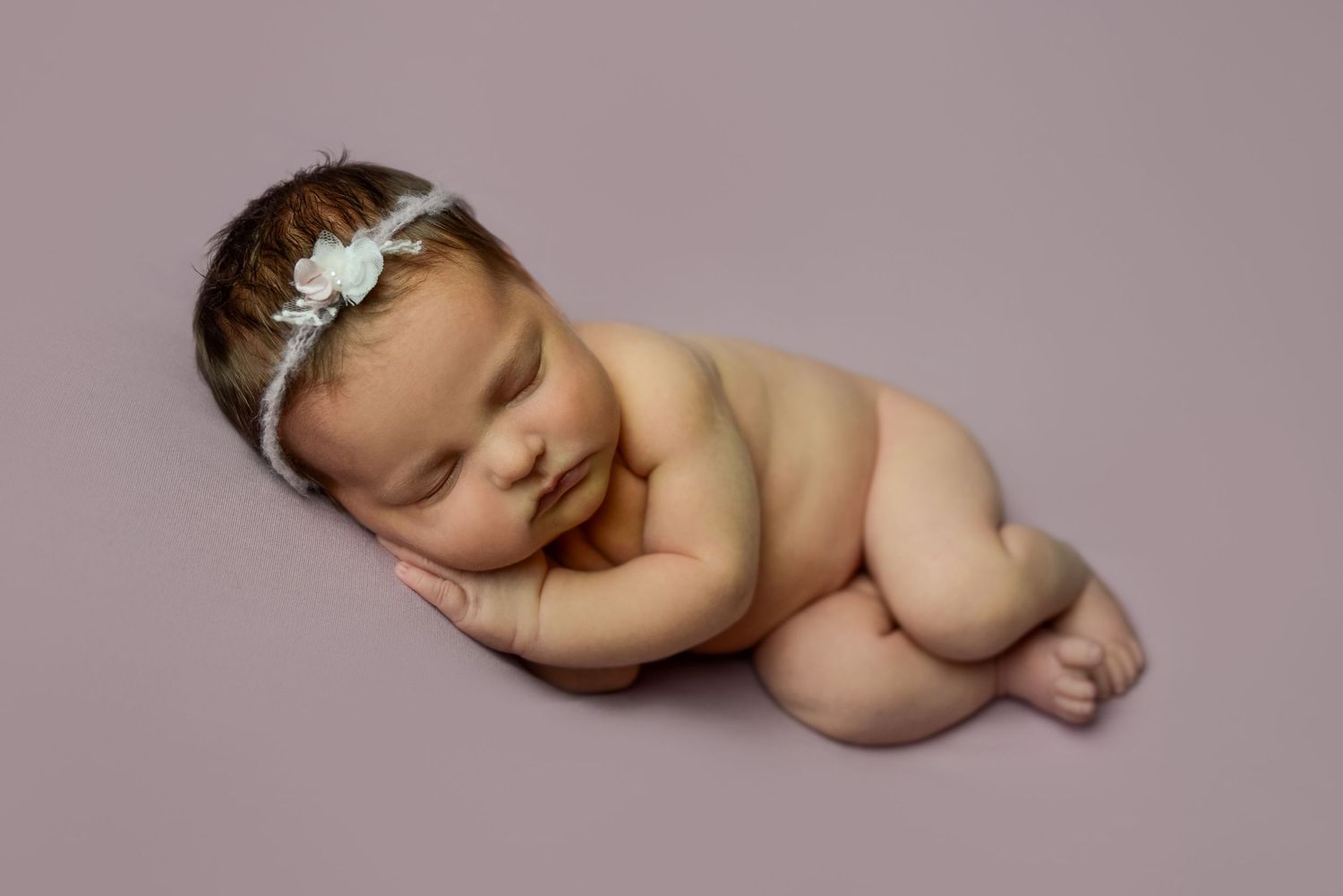Gentle portrait of sleeping baby with white headband against soft pink background.