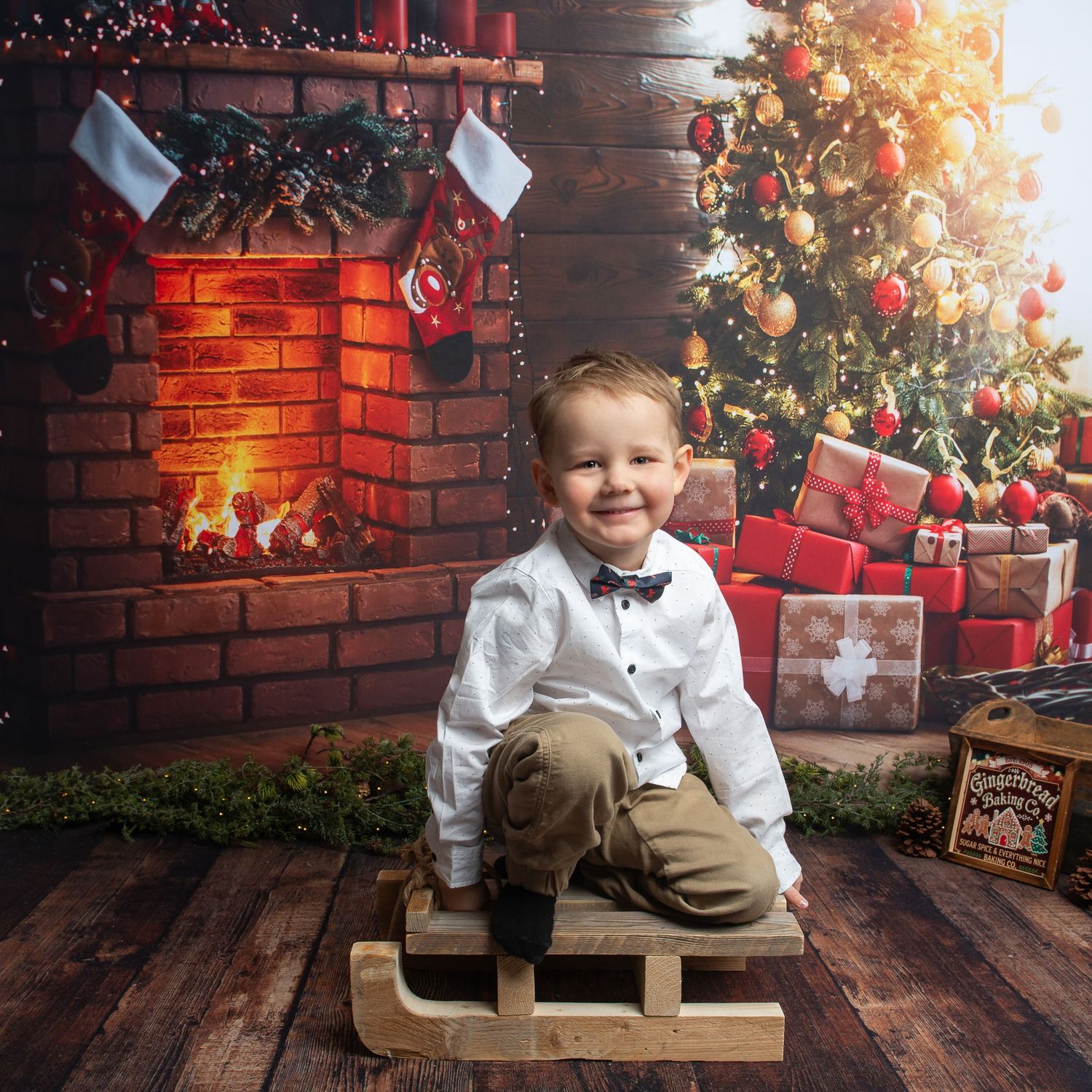 Young boy in white shirt and bow tie sits on a wooden sled before a Christmas tree and lit fireplace.