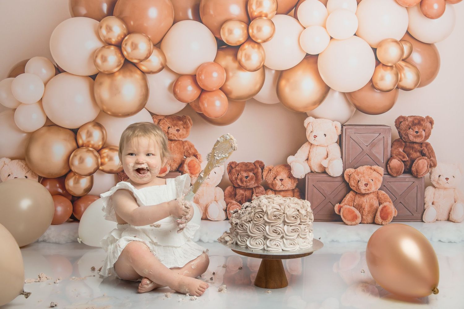 Baby girl smiling at first birthday cake smash with teddy bears and rose gold balloons on a neutral beige backdrop.
