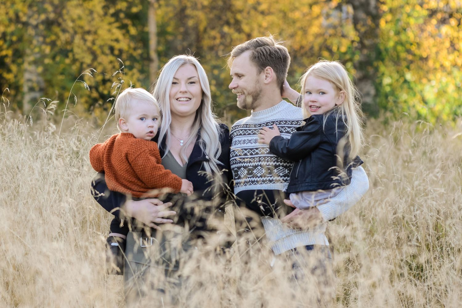 Family of four posing in golden autumn field, parents holding two young children, surrounded by fall foliage.
