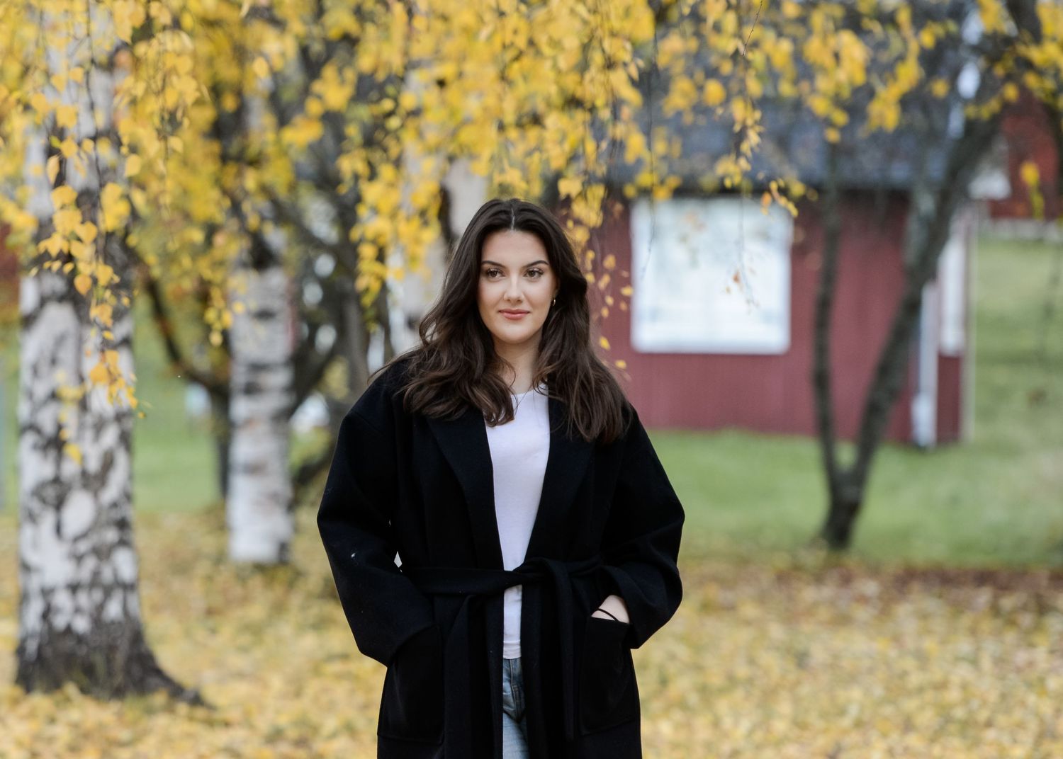 Woman in black coat standing outdoors among birch trees with yellow autumn foliage near a red building.