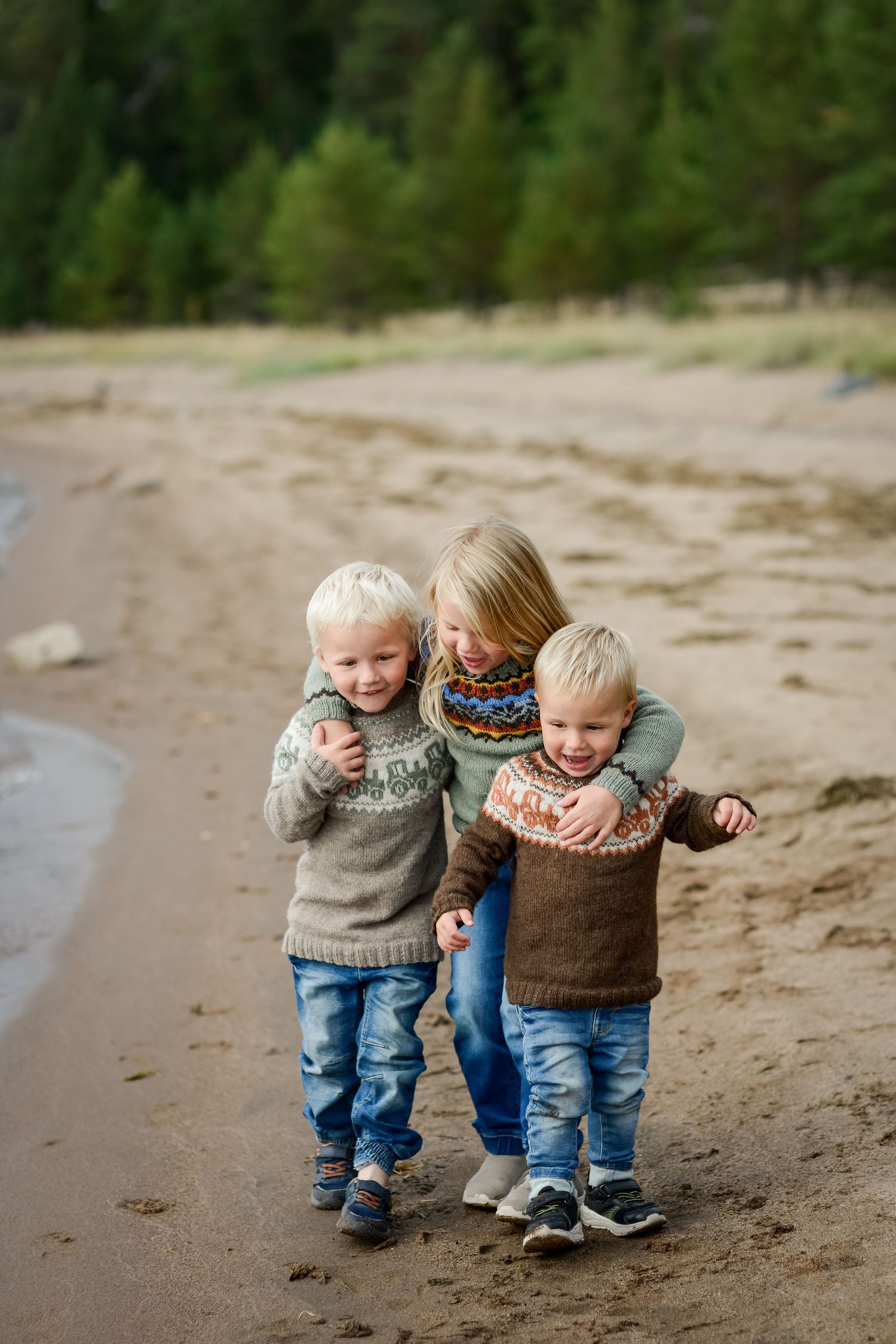 Three blonde children in Nordic sweaters and jeans walk together on a sandy beach with pine trees in background.