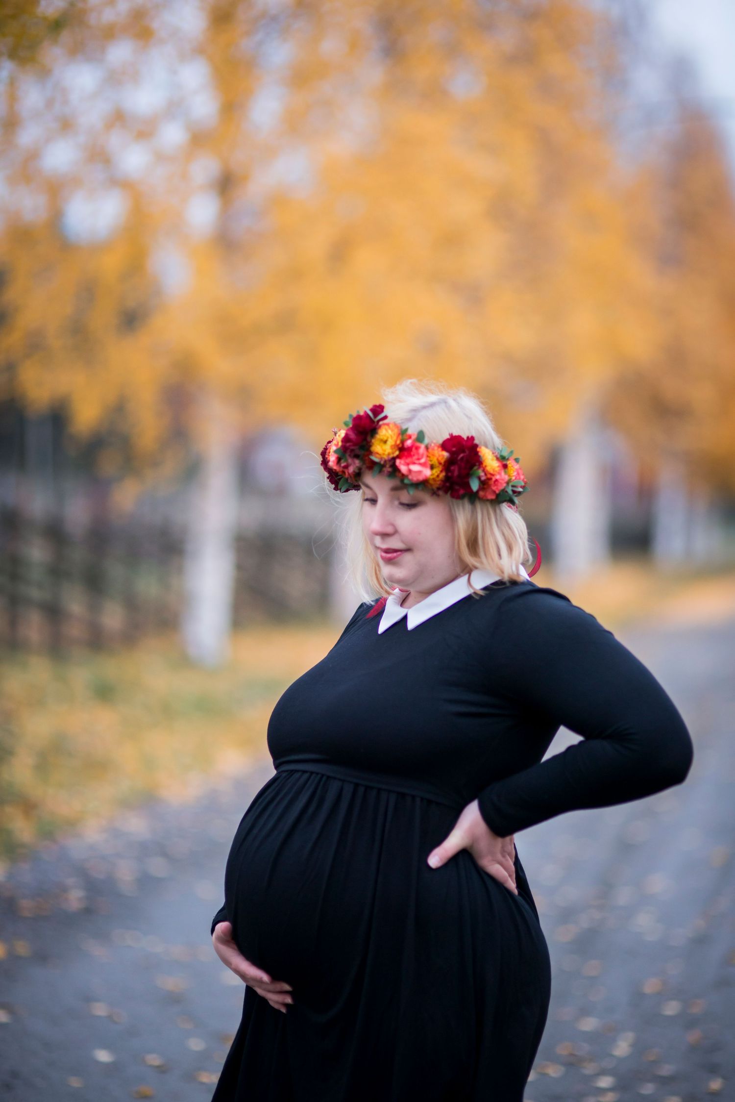 Pregnant woman in black dress and floral crown poses outdoors among golden autumn trees.