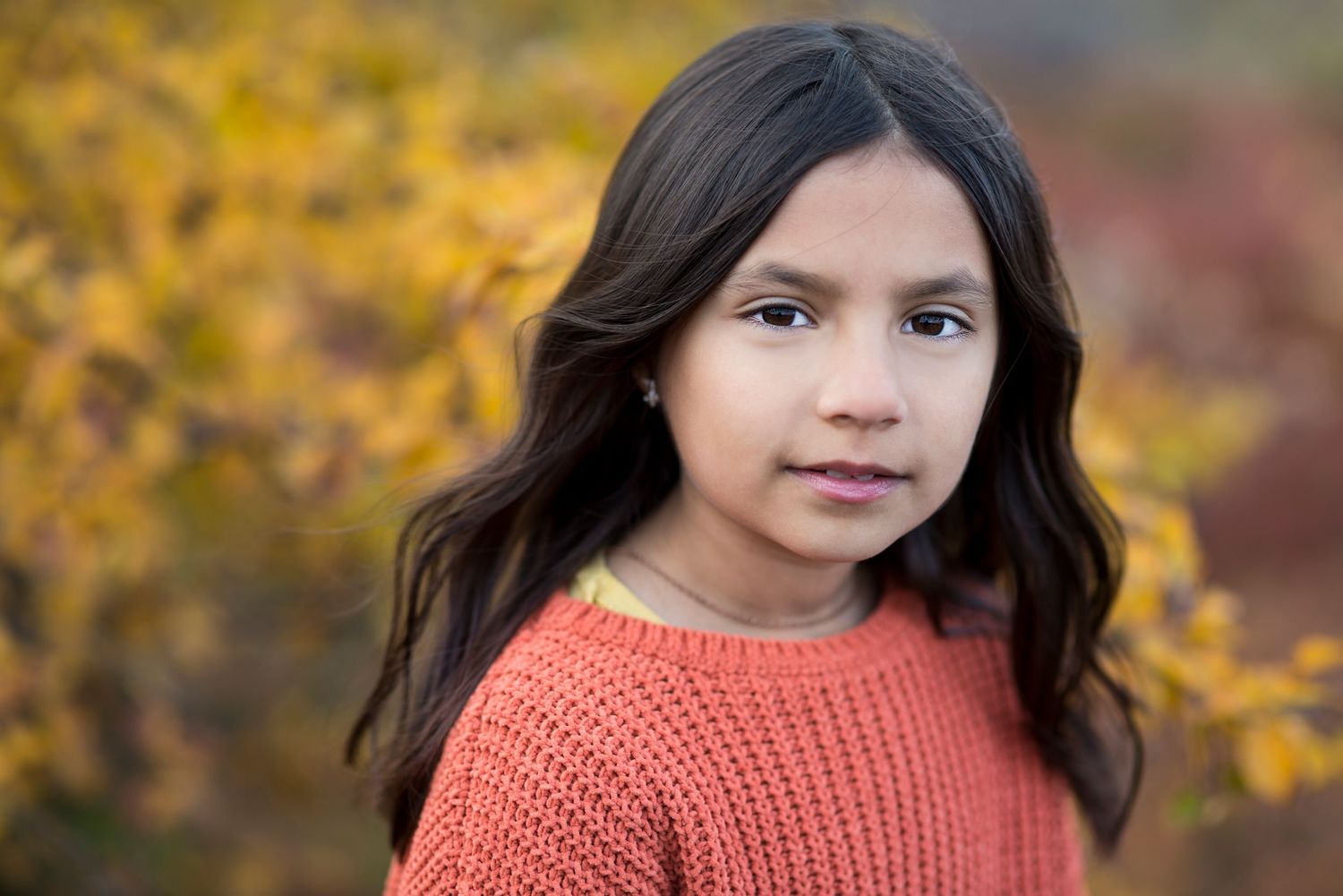 Young girl with long dark hair wearing an orange knit sweater, posing outdoors with autumn foliage background.