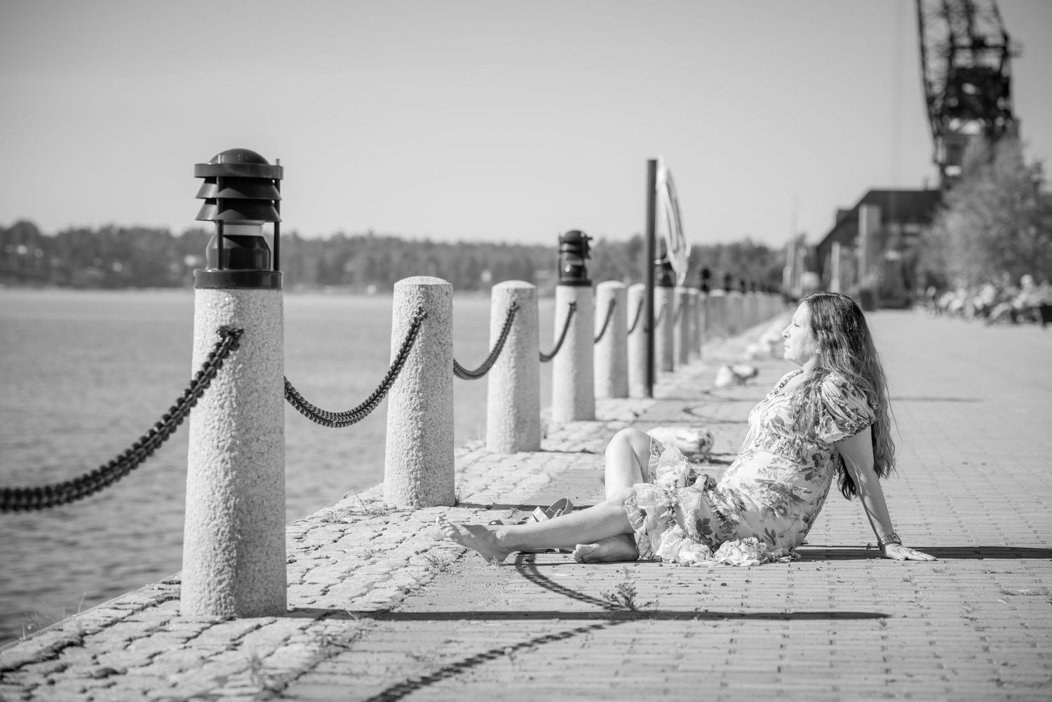 Woman in floral dress sitting on cobblestone waterfront promenade beside rope bollards in black and white.