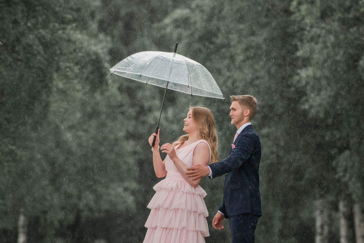 Romantic couple shares an intimate moment under a clear umbrella in a wooded setting with birch trees.