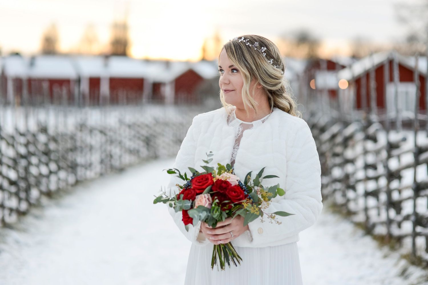 A bride in a white dress stands in fresh snow with red barns visible in the background.