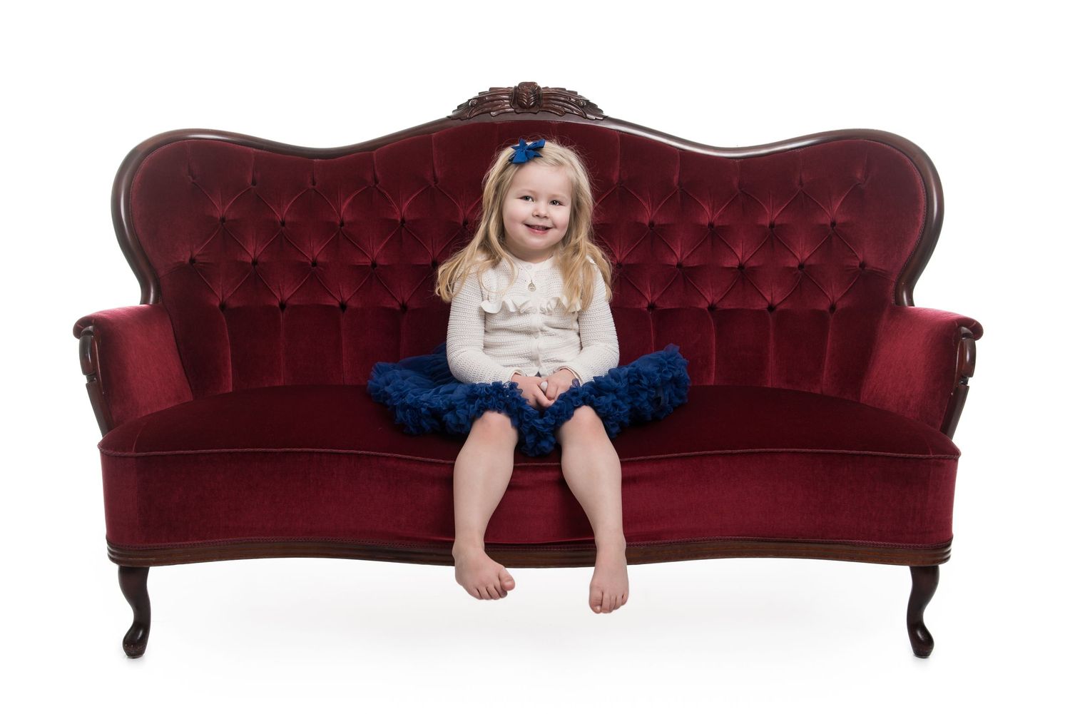 Child in tulle skirt sitting on elegant burgundy tufted vintage sofa.