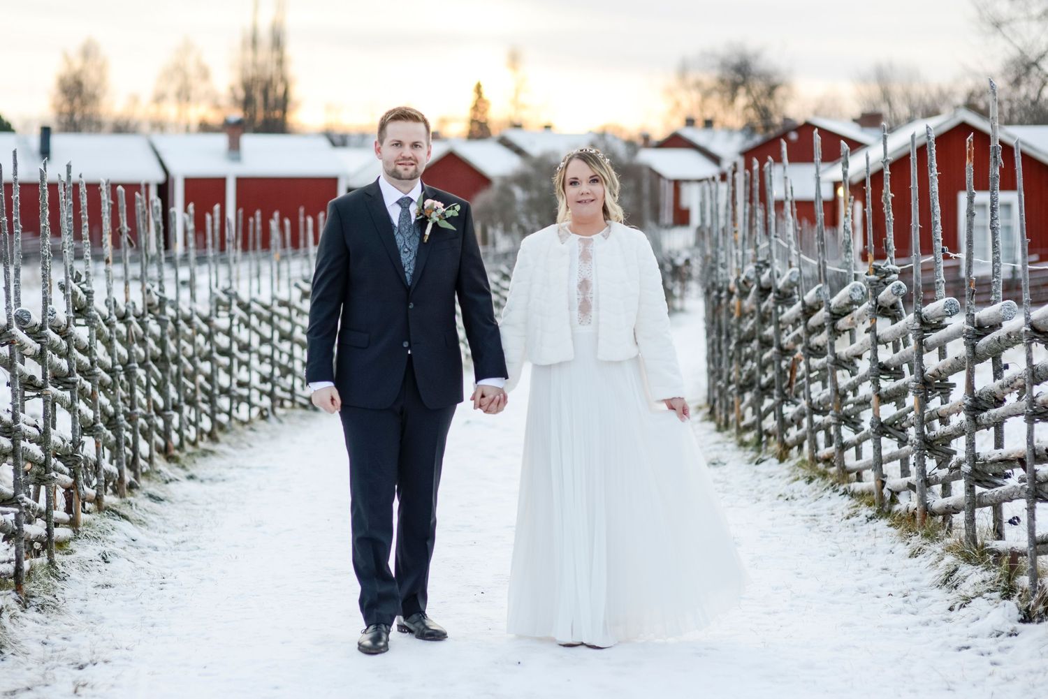 A couple walks hand in hand through snow-covered paths with red buildings in background.