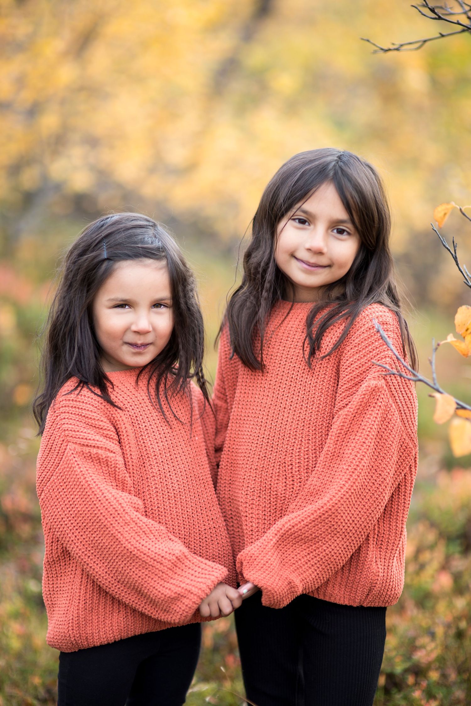 Two young girls in matching coral knit sweaters hold hands while posing together in a vibrant autumn forest setting.