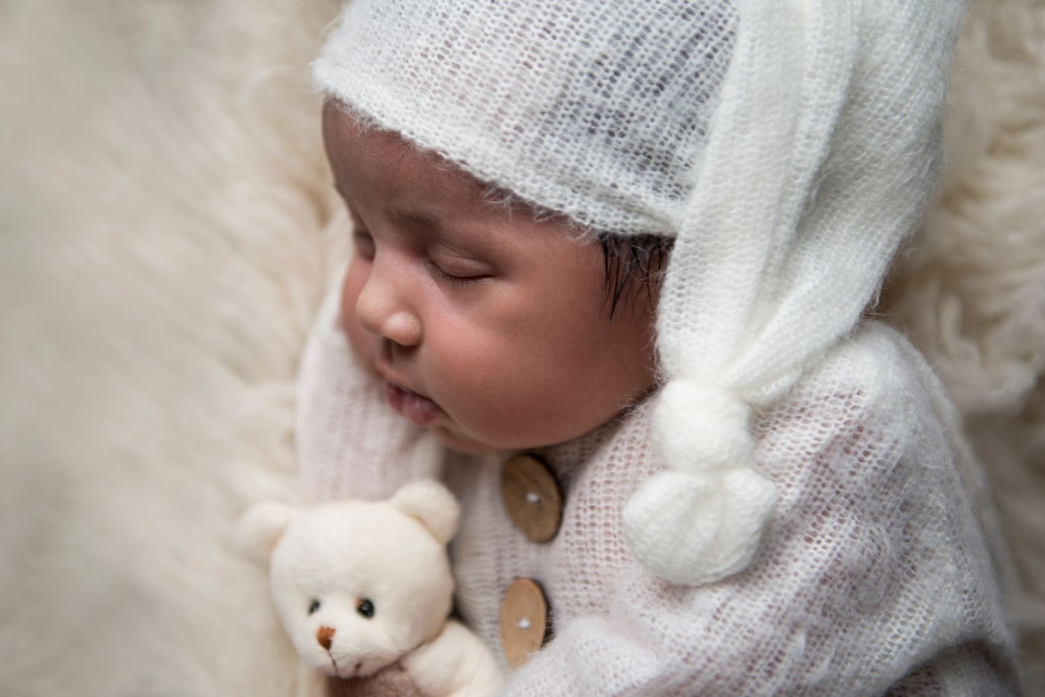Sleeping baby in white knit hat and outfit cuddling small teddy bear on soft backdrop.