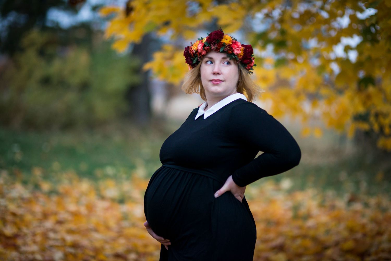 Pregnant woman in black dress and floral crown poses outdoors among autumn leaves in a maternity photo.