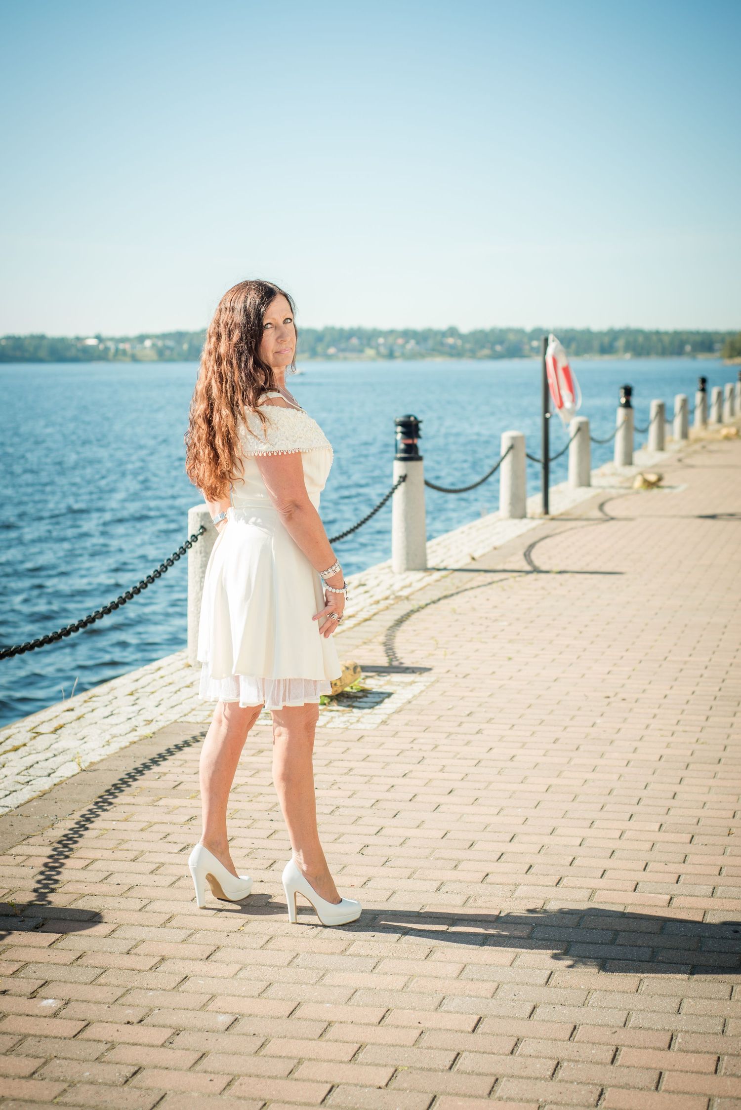 Young woman in white dress and heels poses on a sunny waterfront promenade with blue water in the background.