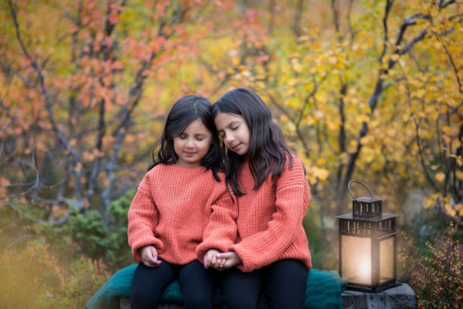 Two young sisters in matching coral sweaters sit together outdoors among colorful autumn foliage with a glowing lantern nearby.