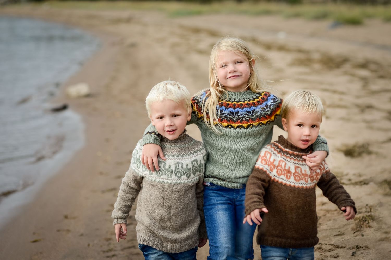 Three children in colorful Nordic sweaters smile on a sandy beach shoreline.