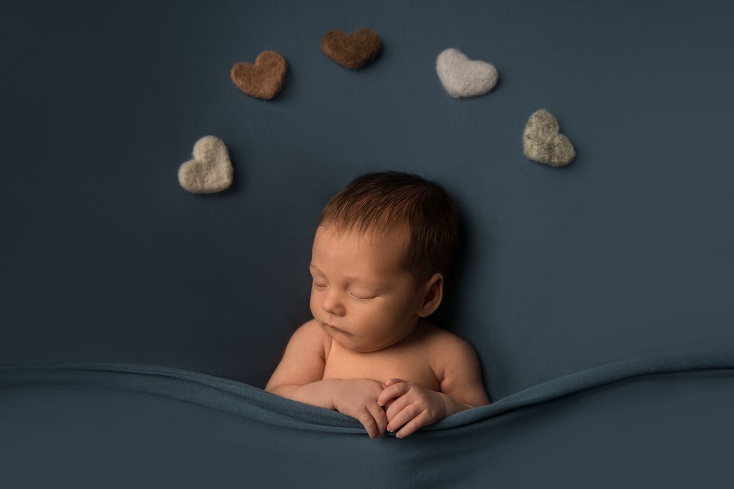 Professional newborn photo series with baby surrounded by heart-shaped props on blue backdrop.