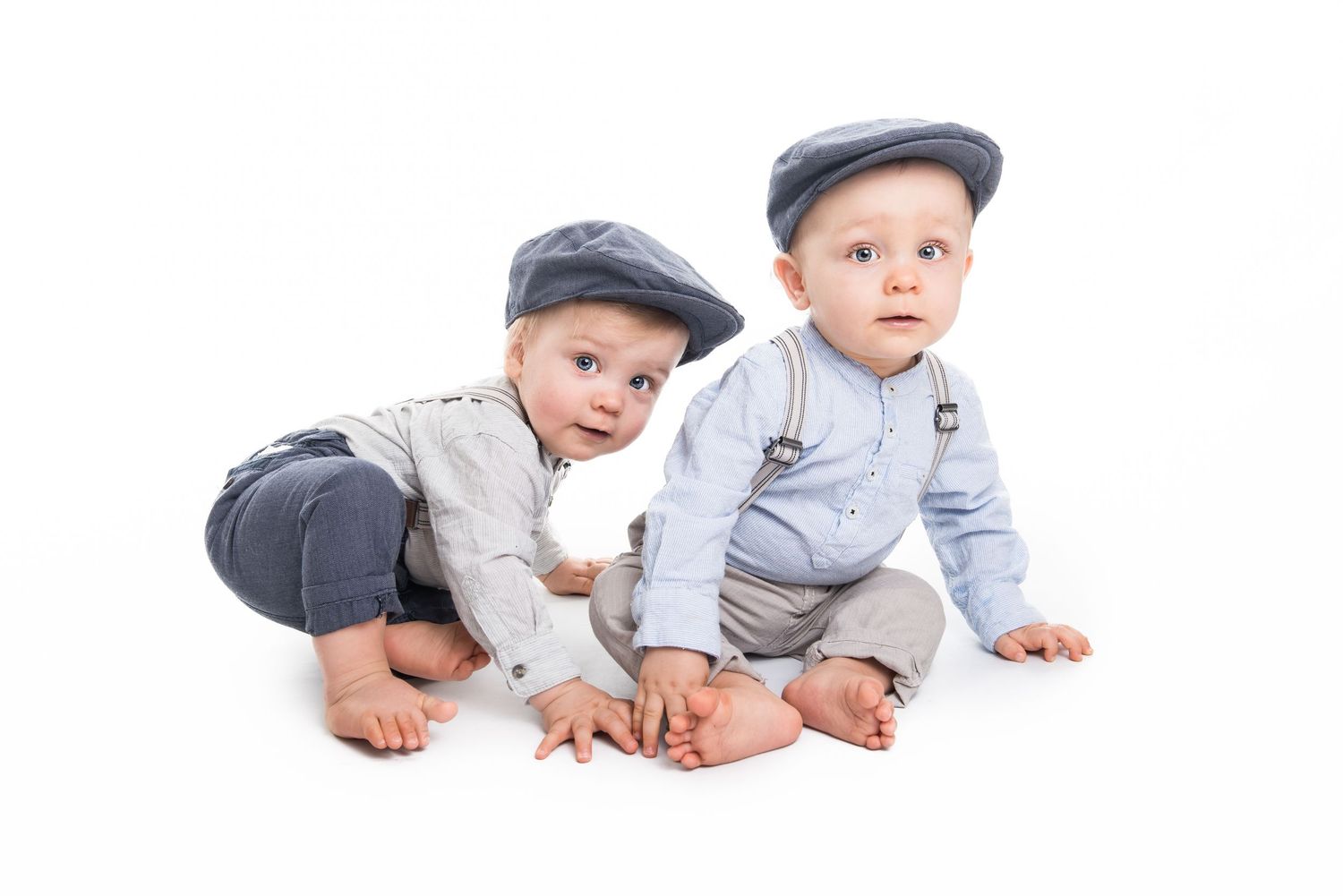 Two toddlers in matching newsboy caps and suspenders sit together on a white background.