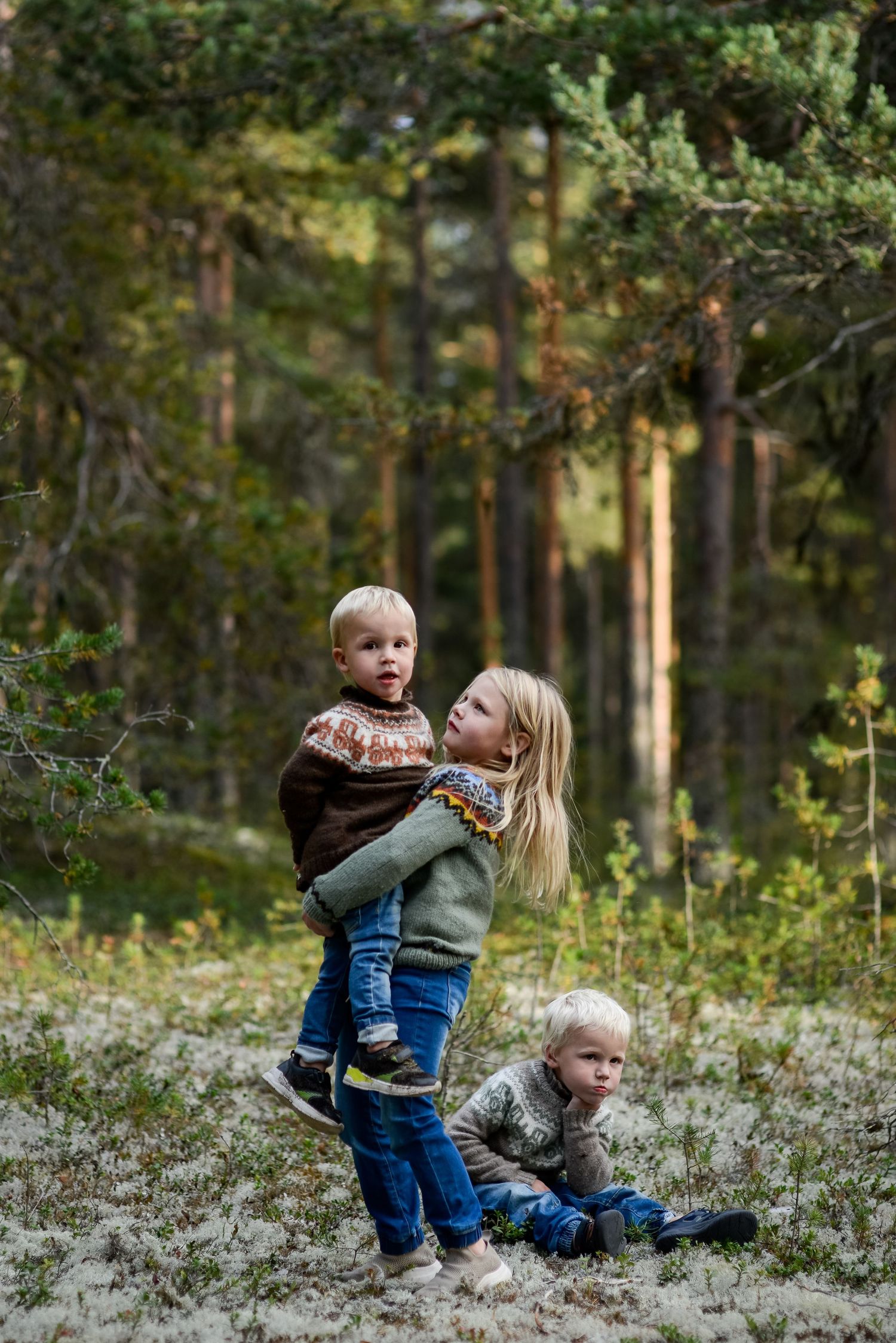 Three young children playing outdoors in a forest, with a girl holding a toddler while another child sits on the ground.