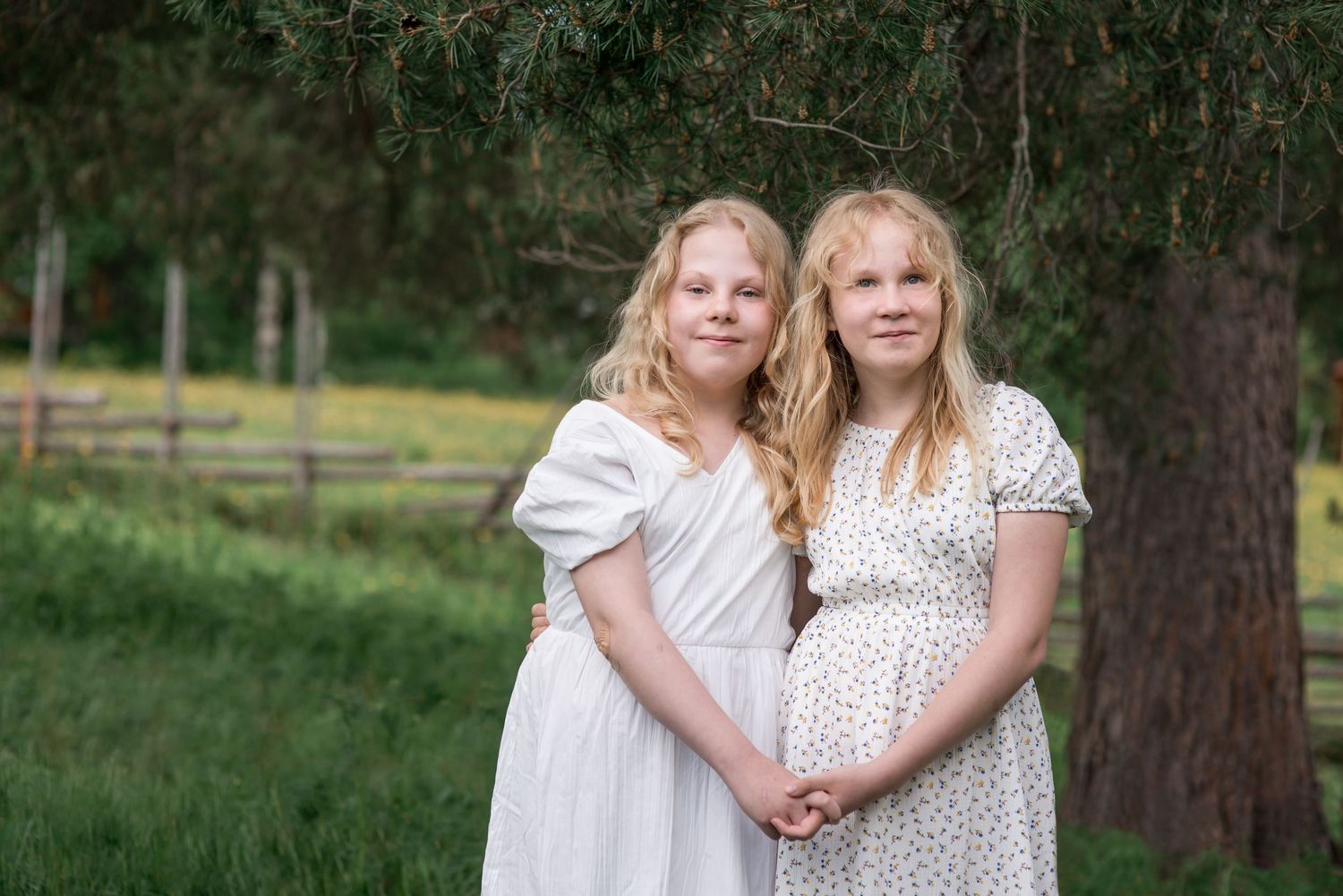 Two blonde girls in white dresses pose together outdoors near pine trees and a wooden fence.
