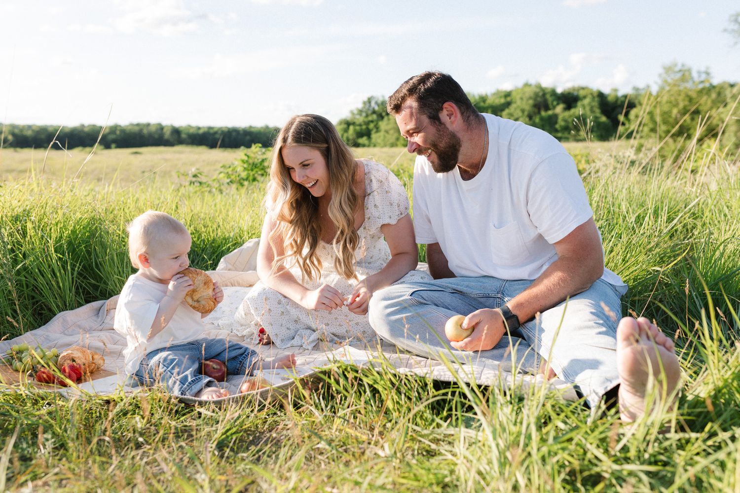 Young family enjoying a sunny outdoor picnic on a blanket in a green field with their baby.