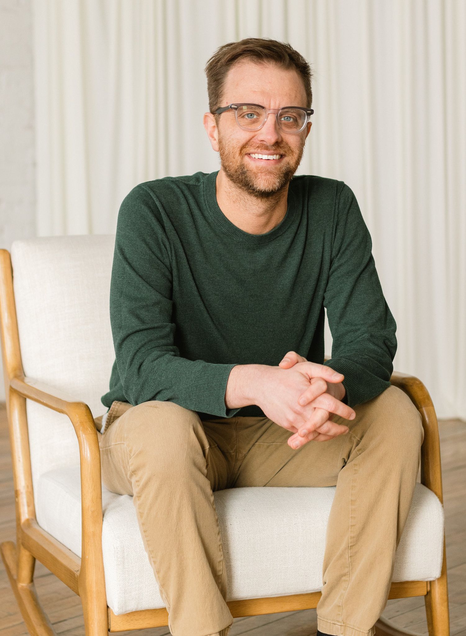 Smiling man with glasses in green sweater and khaki pants sitting relaxed on white chair in bright modern room.