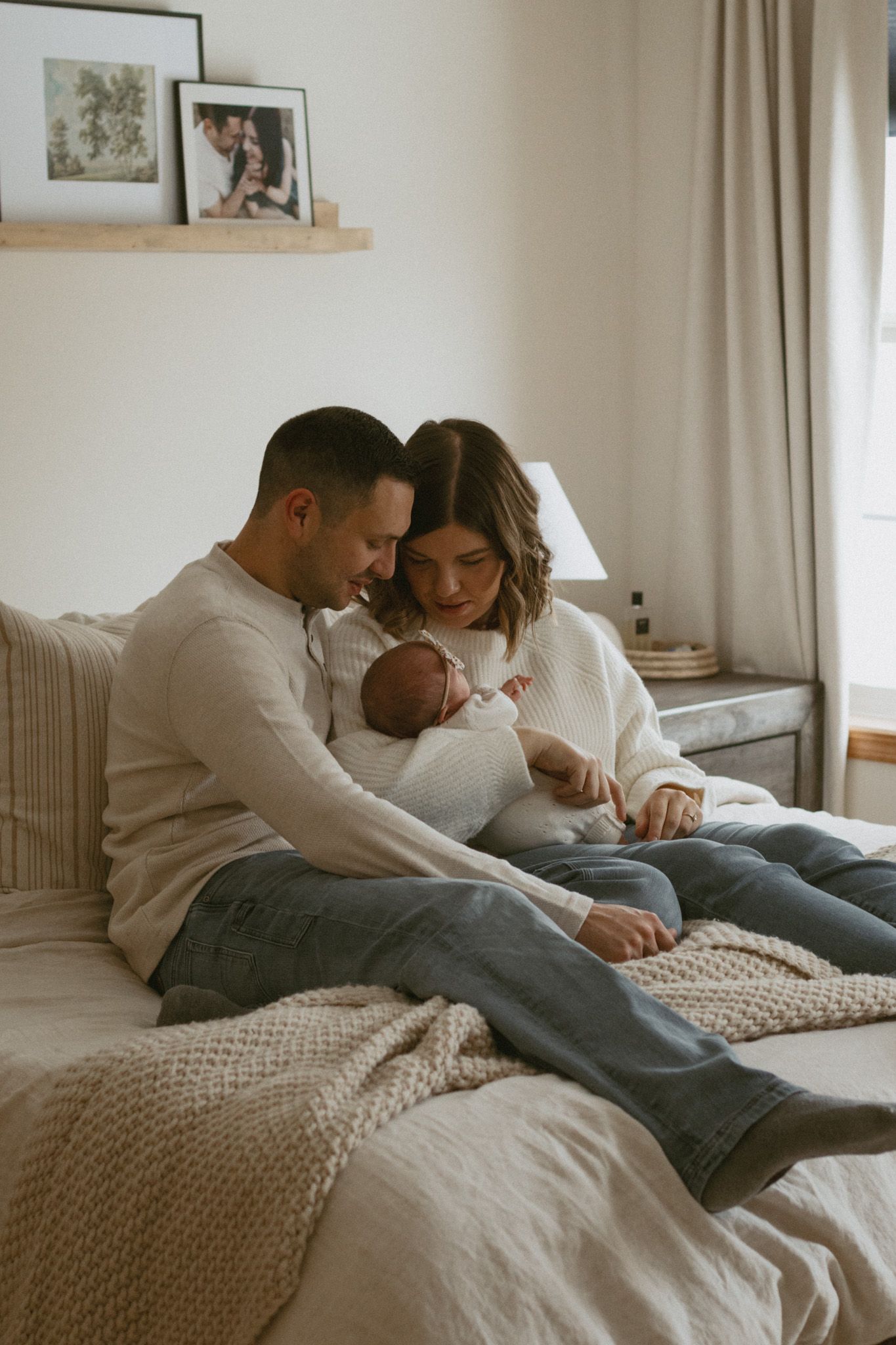 A couple sits on a bed gazing lovingly at their newborn baby in a cozy, softly lit bedroom in Otsego, MN.