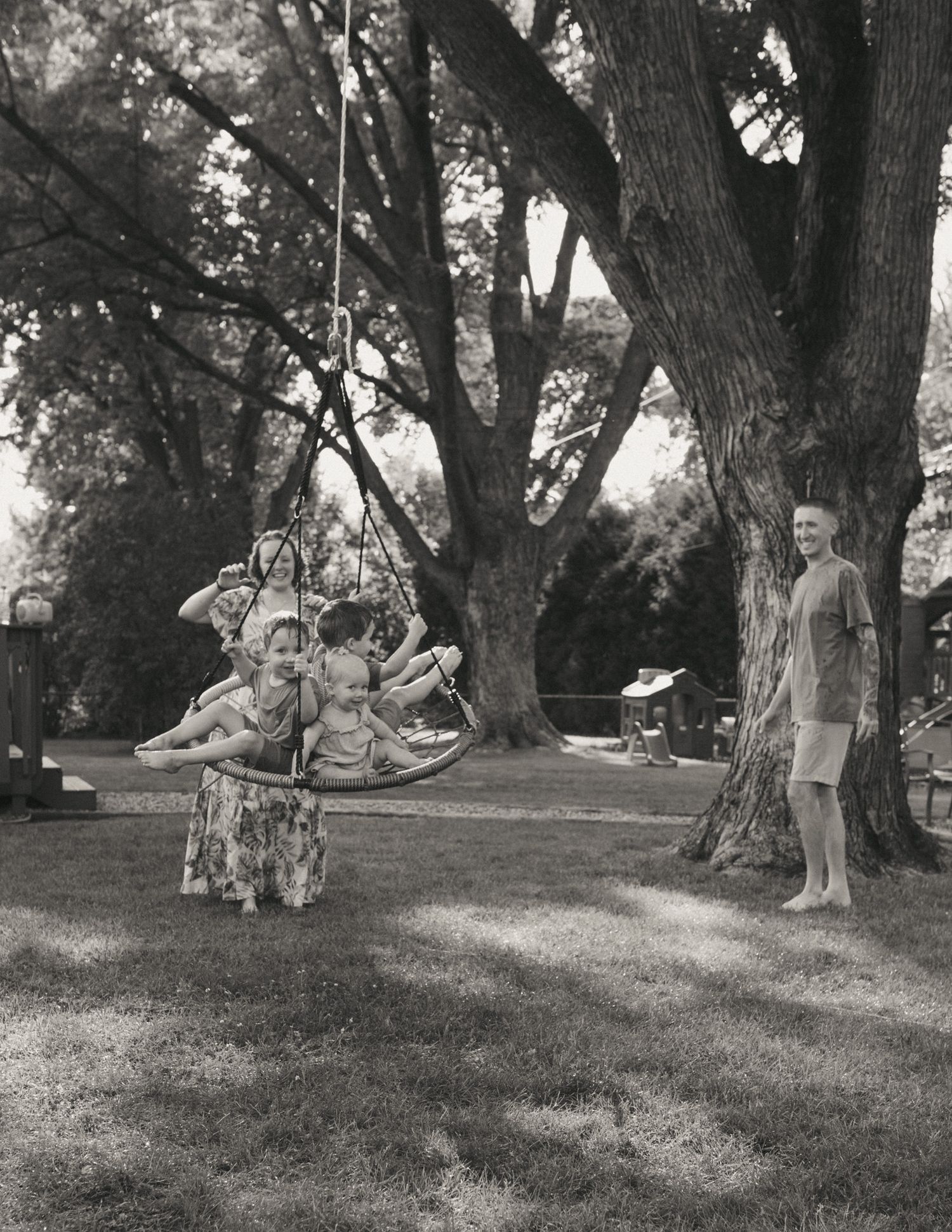 Black and white photo of children playing on a rope swing under large trees while an adult watches nearby.