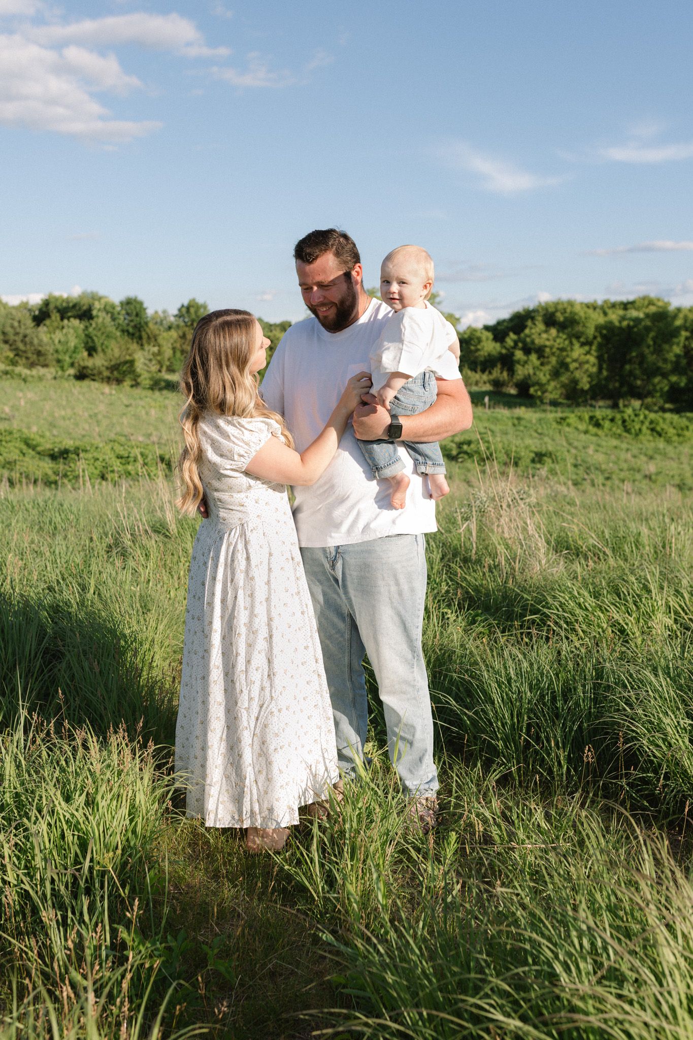 A young family with a baby stands in a lush green field under a blue sky on a sunny day.