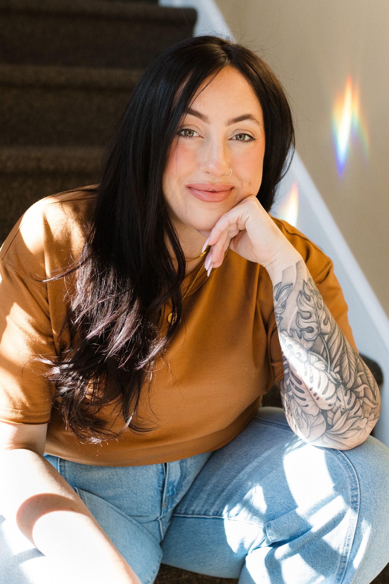 Female Mental health therapist with black hair and sleeve tattoo sitting on stairs in brown shirt and jeans, smiling in St. Paul.