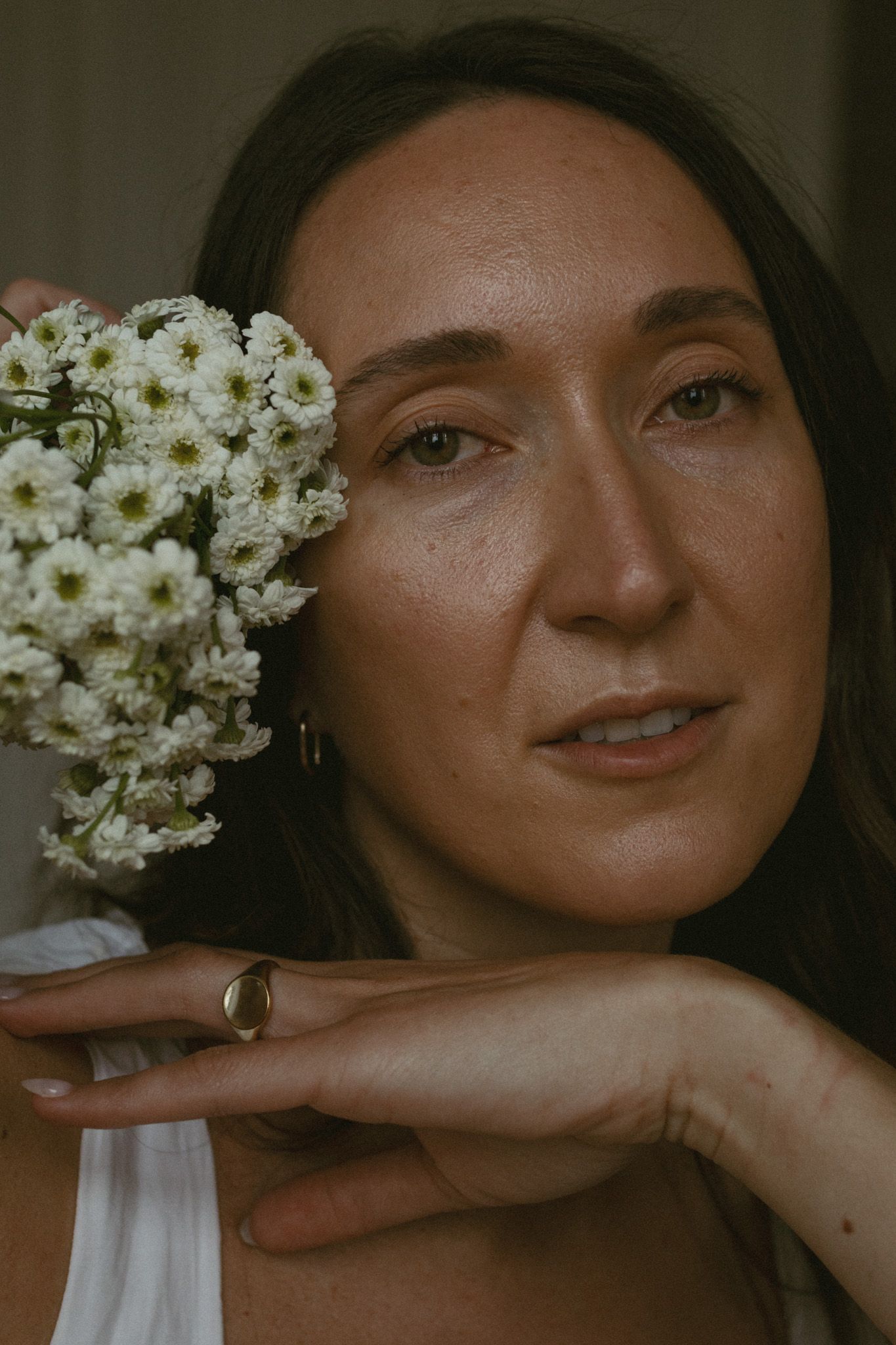 Dark-haired woman holding white chrysanthemum flowers beside her face, wearing gold ring, soft lighting.