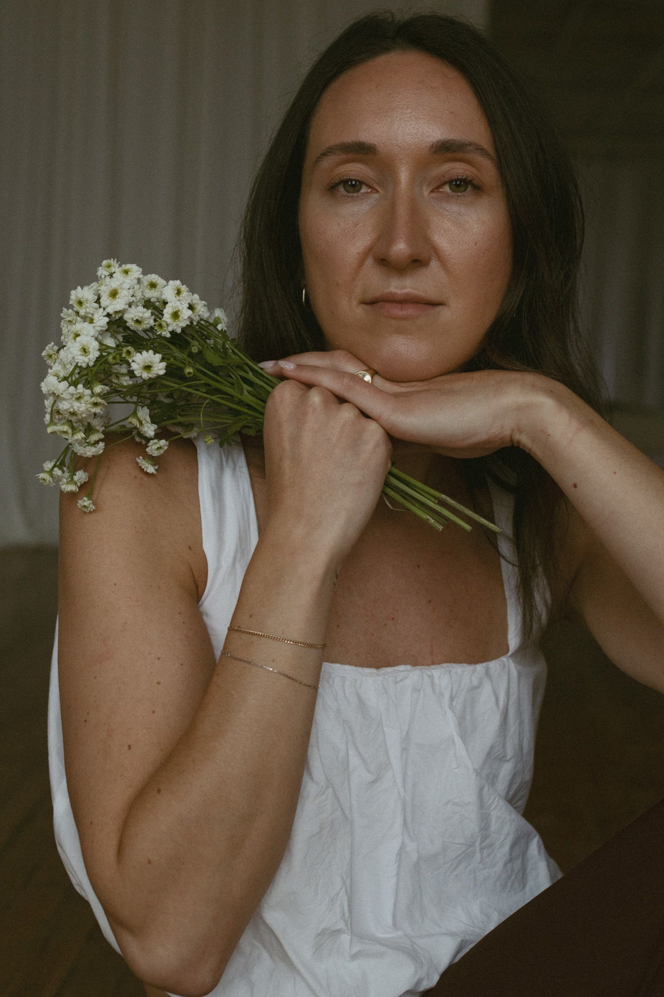 Woman in white top holding white daisy bouquet, posing thoughtfully with hand under chin indoors.