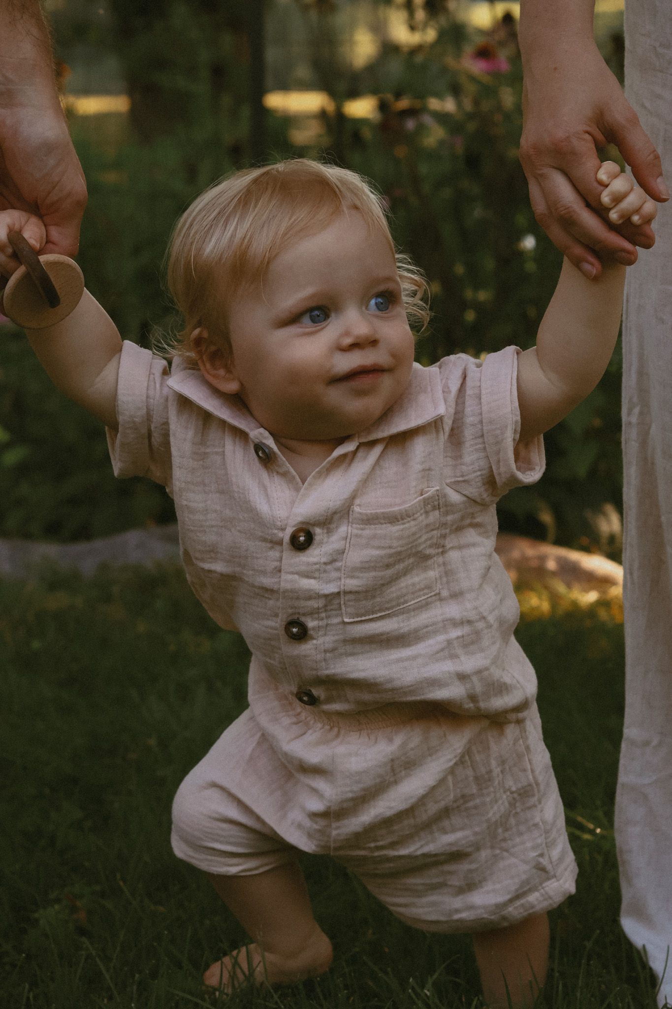 Adorable blonde toddler in a linen outfit taking first steps while holding adults hands in a garden.