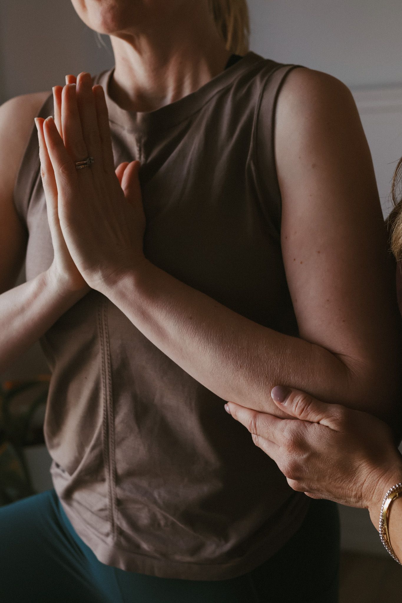 Person in brown tank top practicing yoga prayer hands pose with instructor guiding alignment.